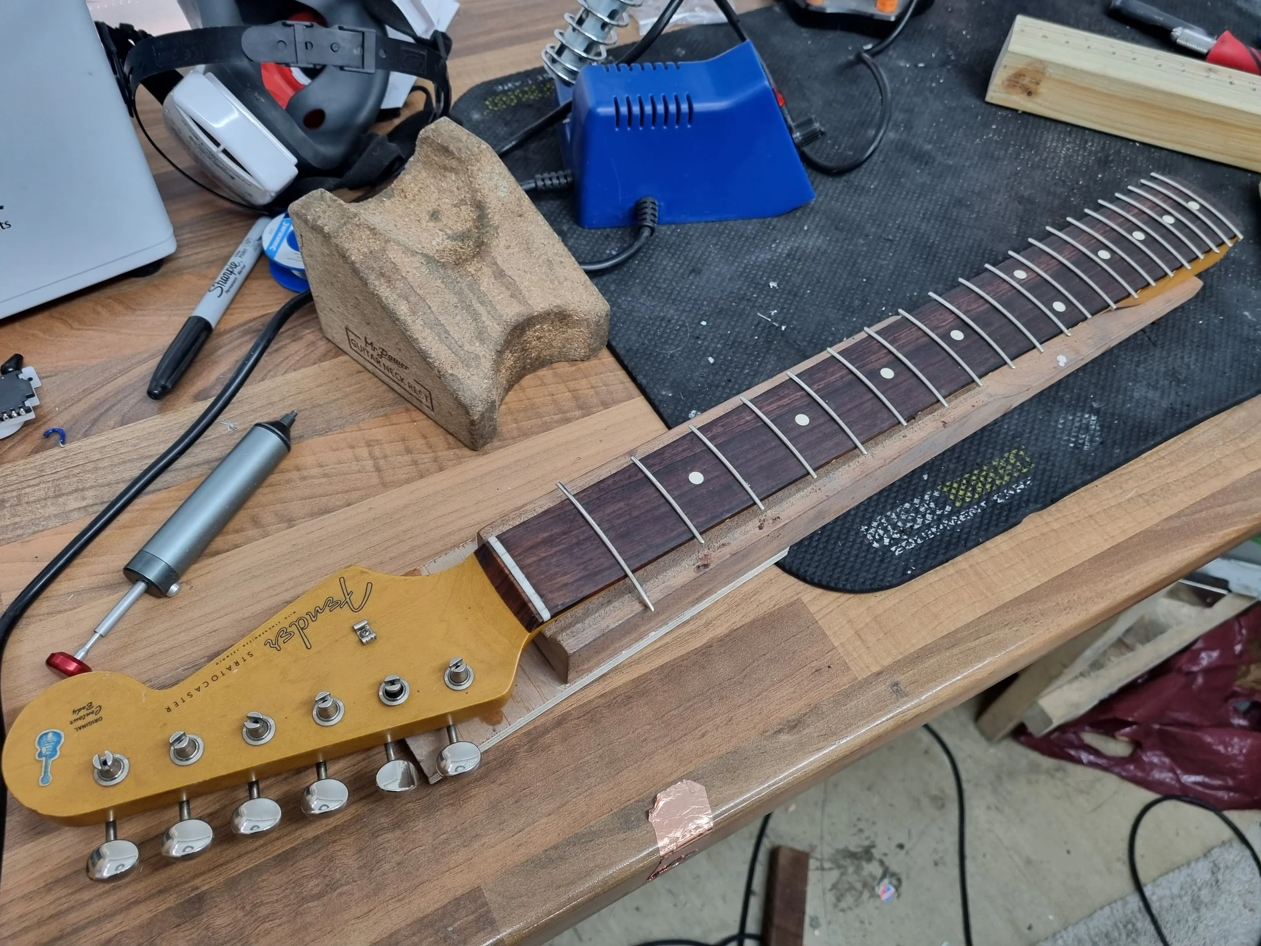 A guitar neck being refretted, placed on a workbench with tools, a workpiece, and a heat gun nearby, indicating a guitar repair or construction process.