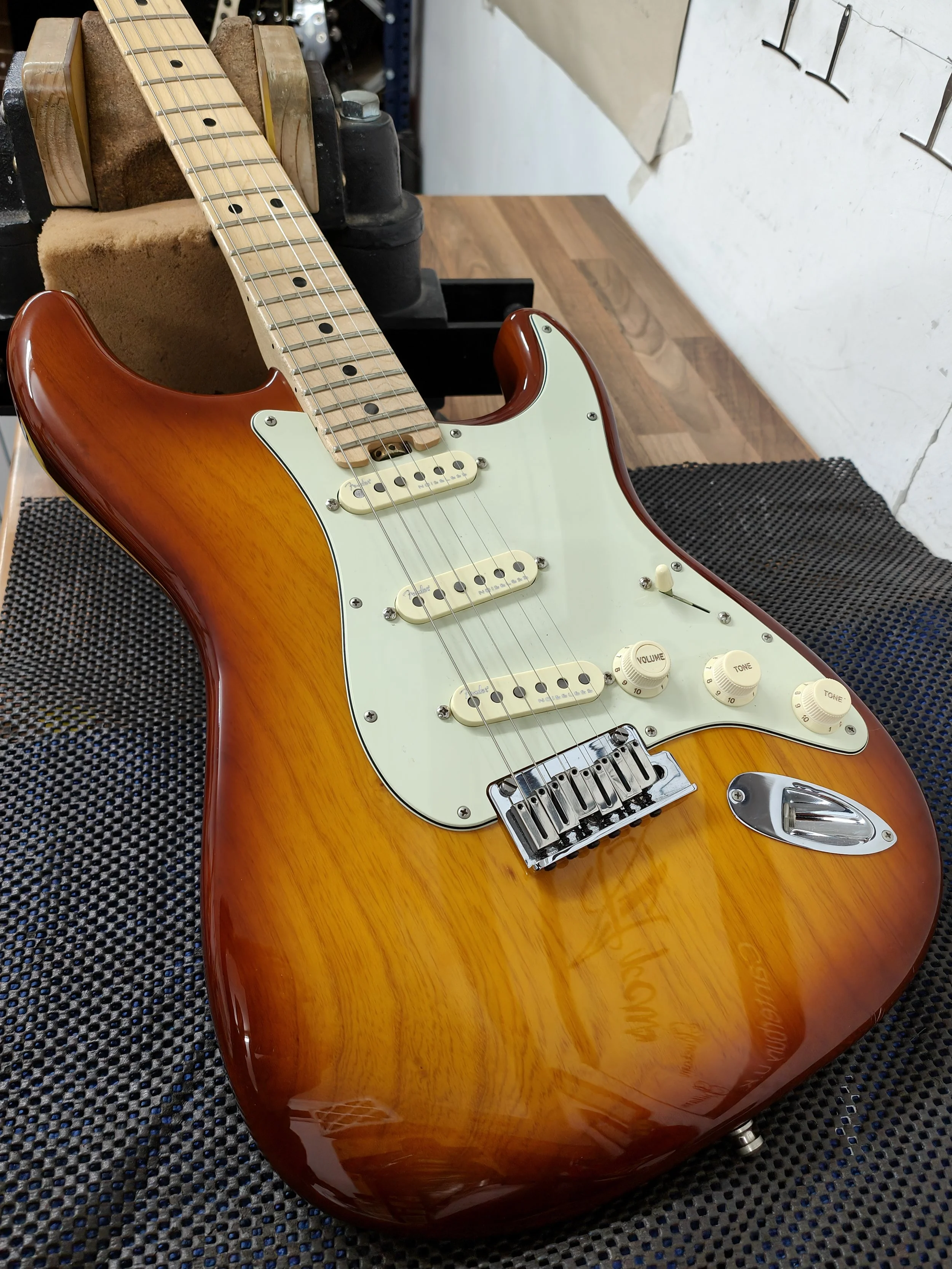 An electric strat guitar with a sunburst finish, maple neck, and white pickguard on a workbench.