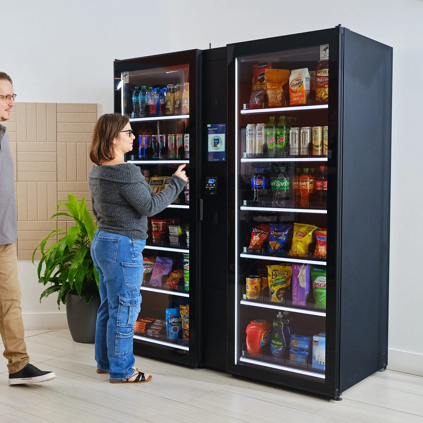 Two people using smart ai cooler vending machines by Petite Cafe with glass doors filled with snacks inside.
