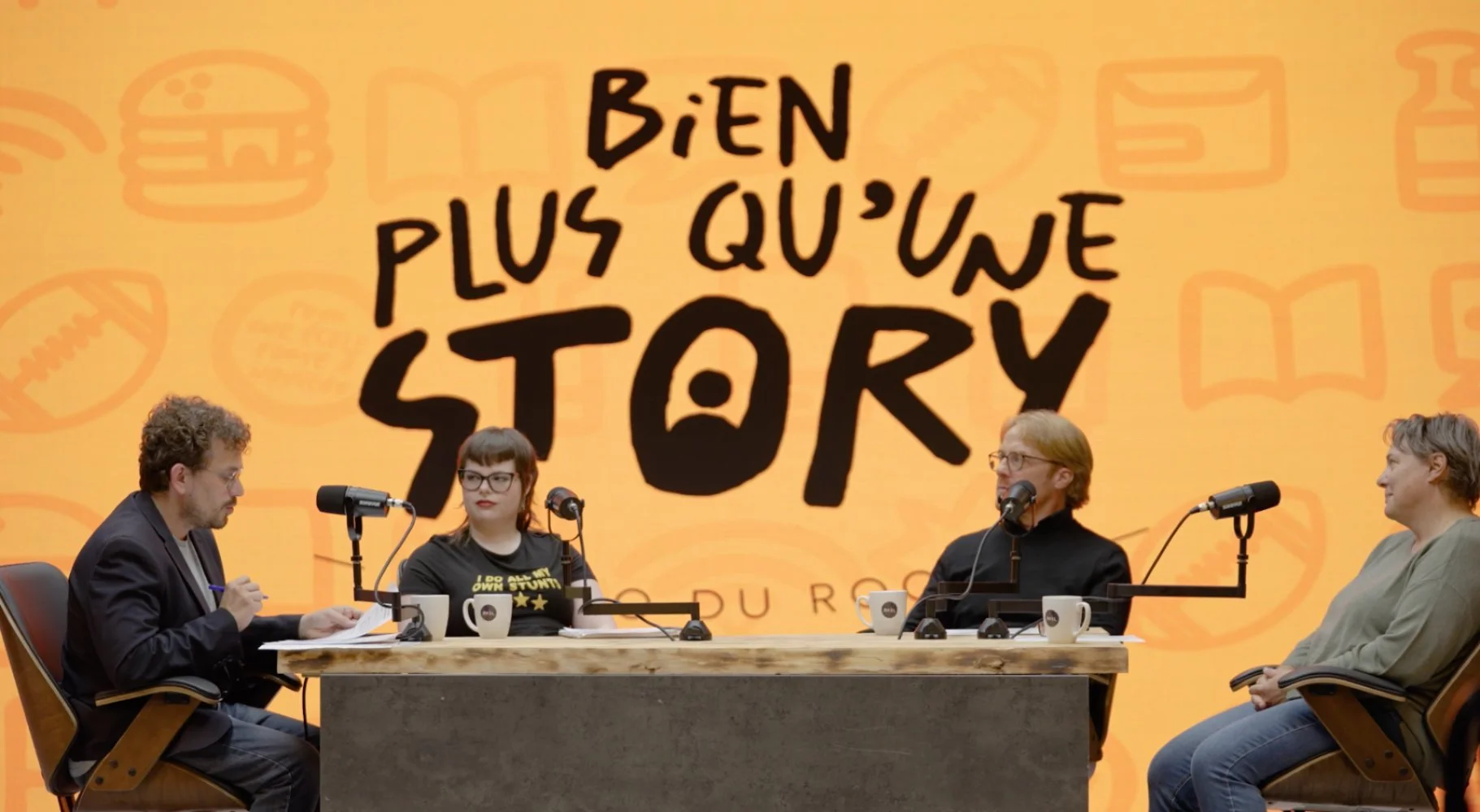 Four people sitting at a table during a panel discussion, with a large orange backdrop displaying the message "Bien plus qu'une story" and various icons in the background.