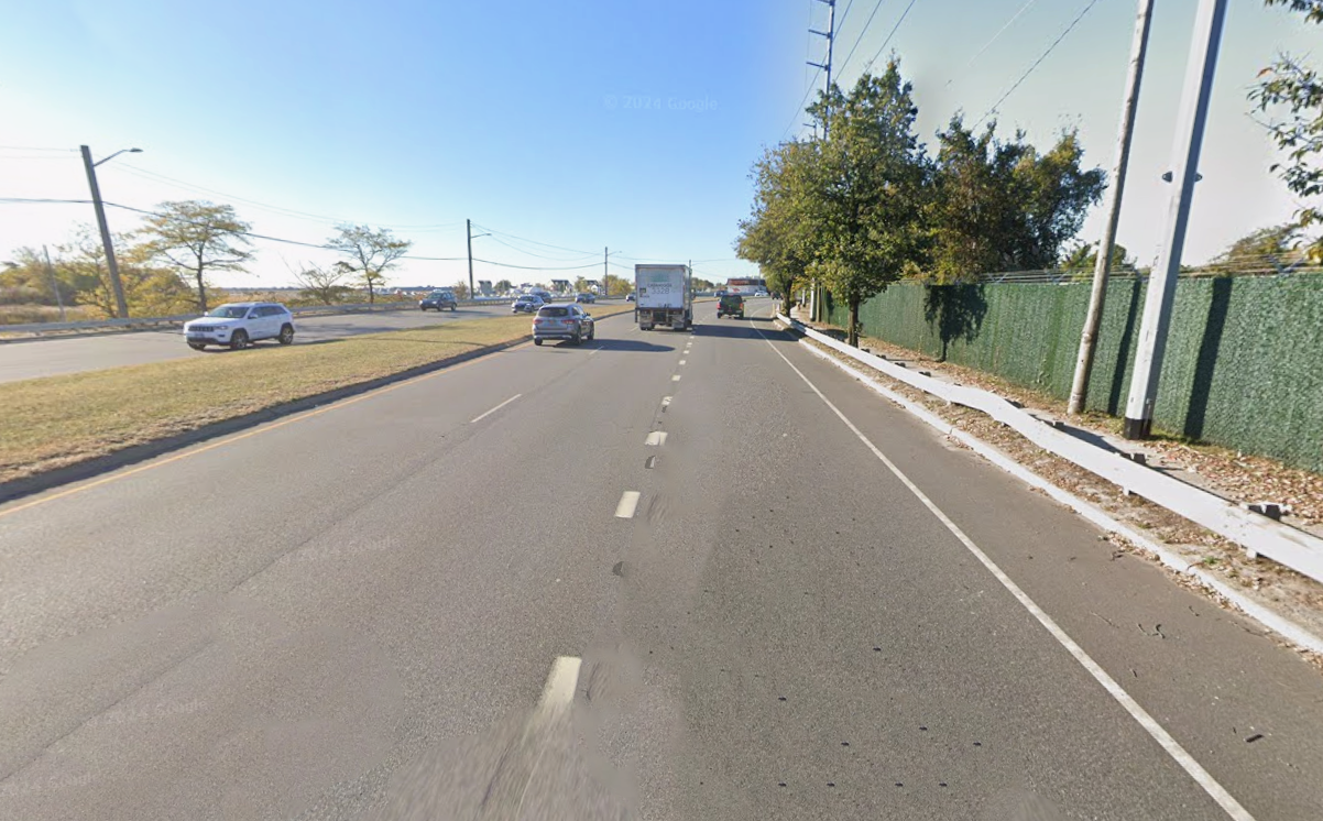 A street view showing traffic with cars and a truck driving, a green fence on the right side, and trees in the background under a clear blue sky.