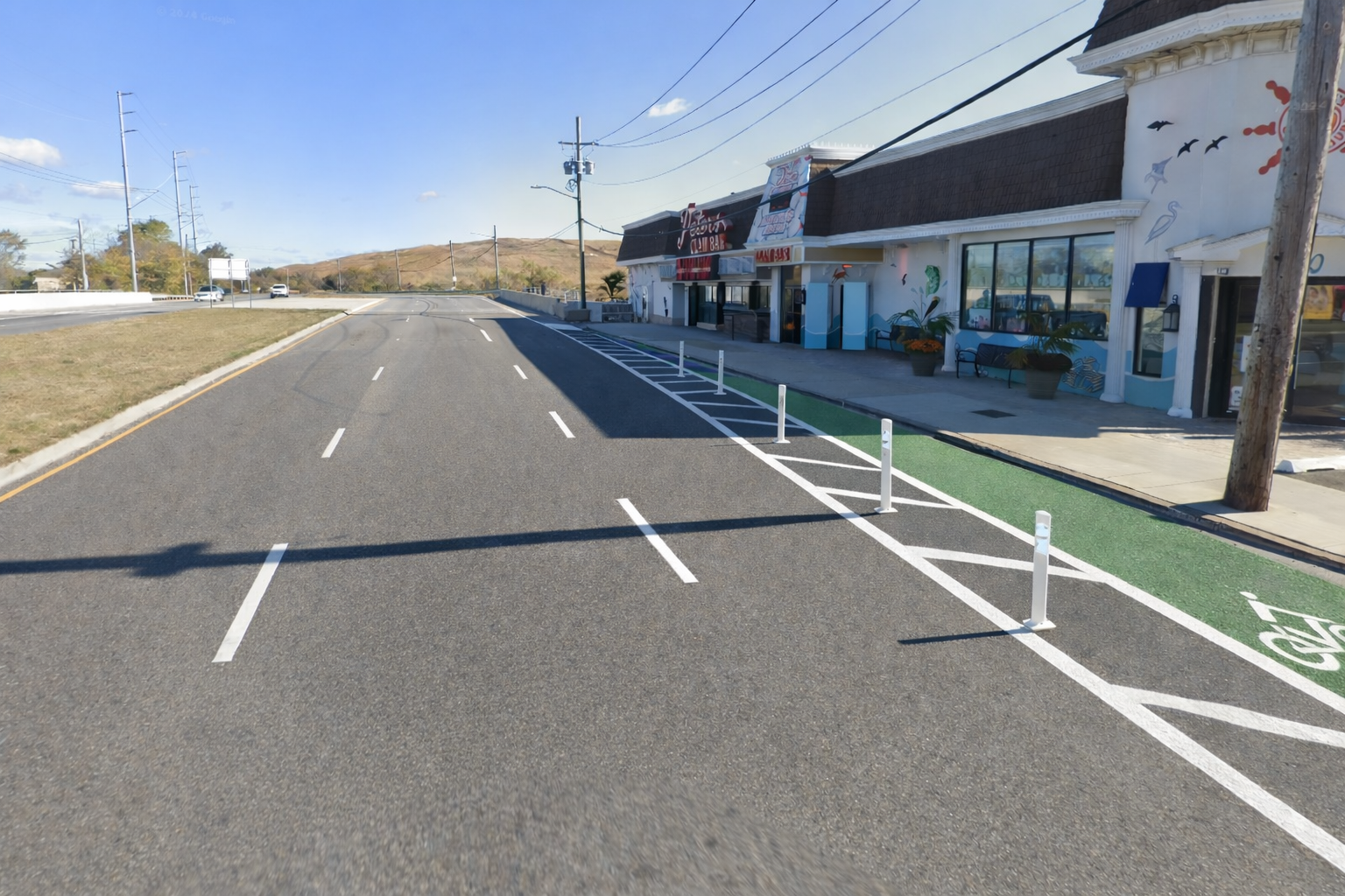 Empty street with parking spaces, a building with storefronts, painted murals, and a mountain in the background under a partly cloudy sky.
