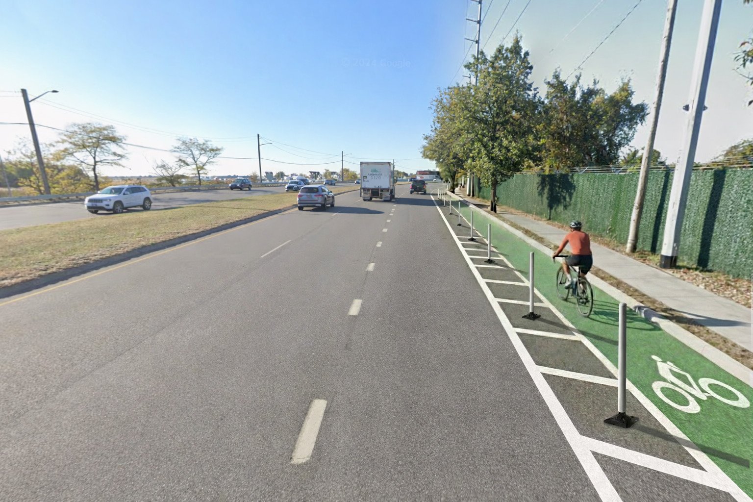 A road with a bike lane on the right side, separated by green paint and poles. A cyclist wearing a helmet and orange shirt is riding on the bike lane. Vehicles are traveling on the main road on the left side, and trees line the sidewalk on the right.