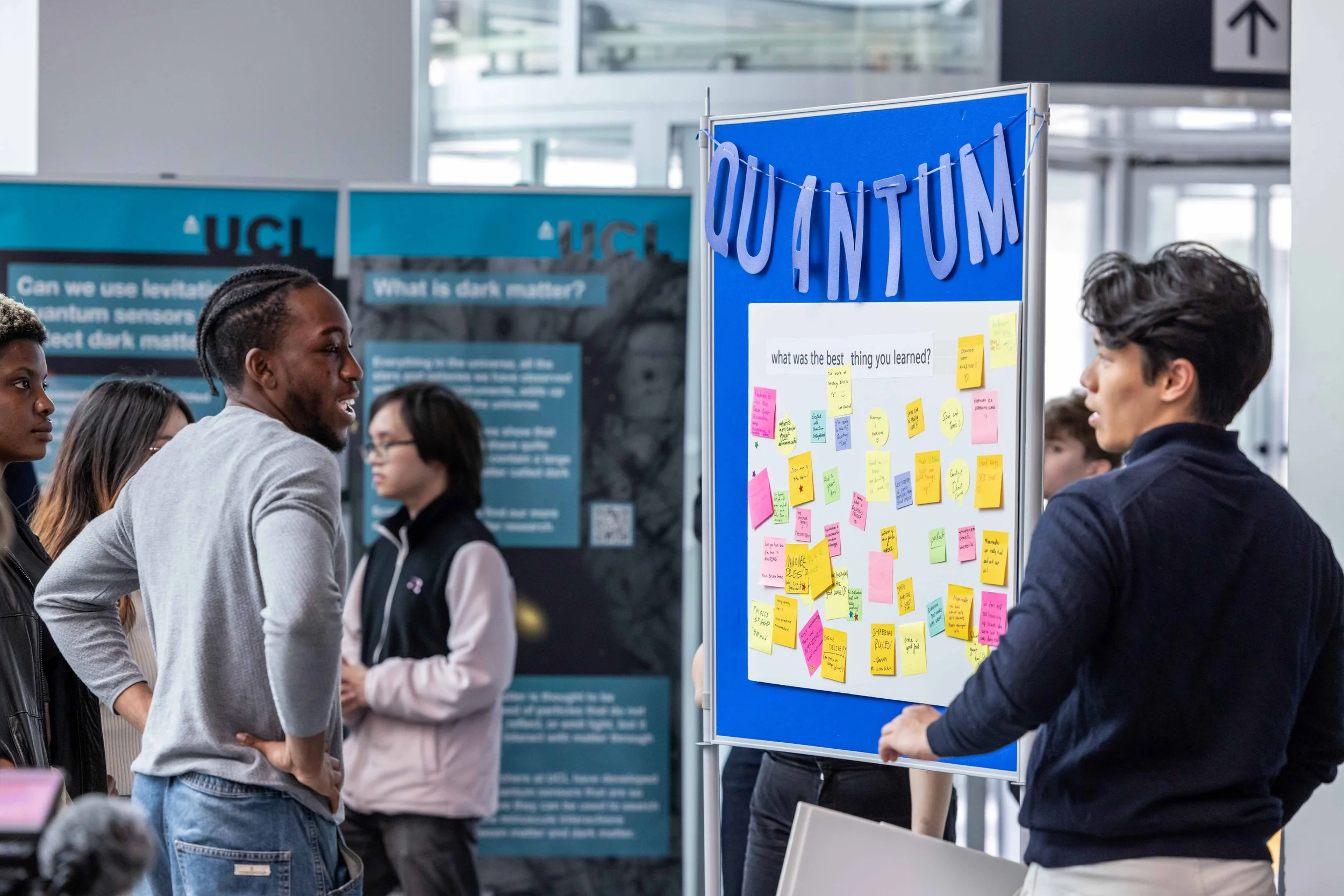 Students at a science fair presenting project on quantum physics with a poster decorated with colorful sticky notes and a sign that says 'QUANTUM'.