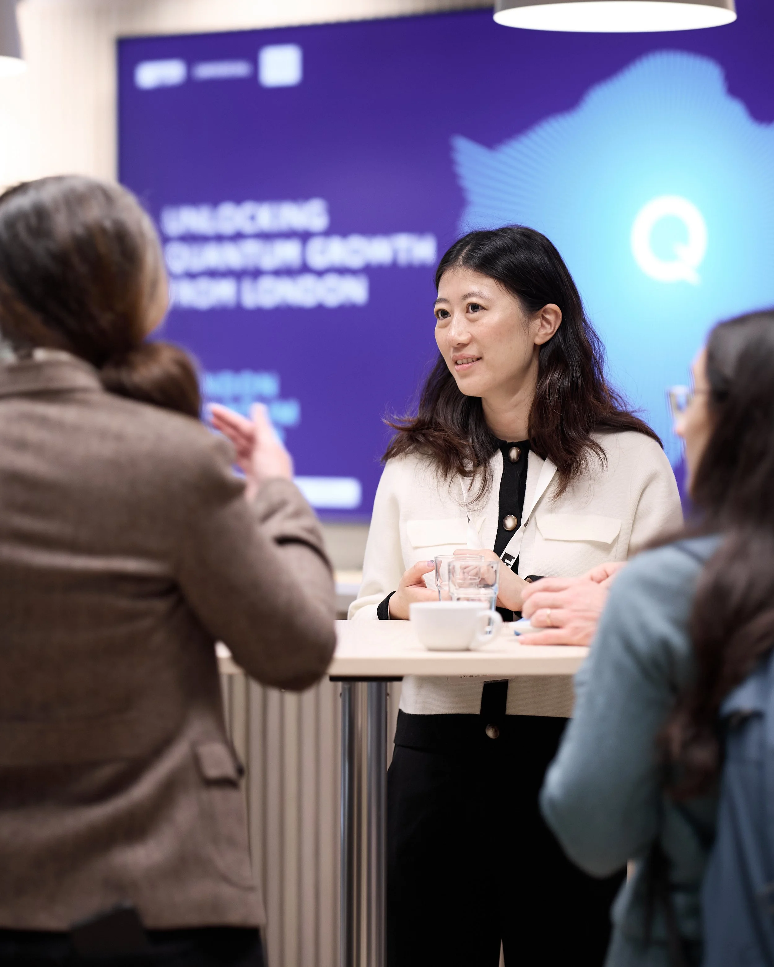 A woman in a white blazer engages in a conversation with two others at a round table, with a large blue digital screen in the background displaying text.