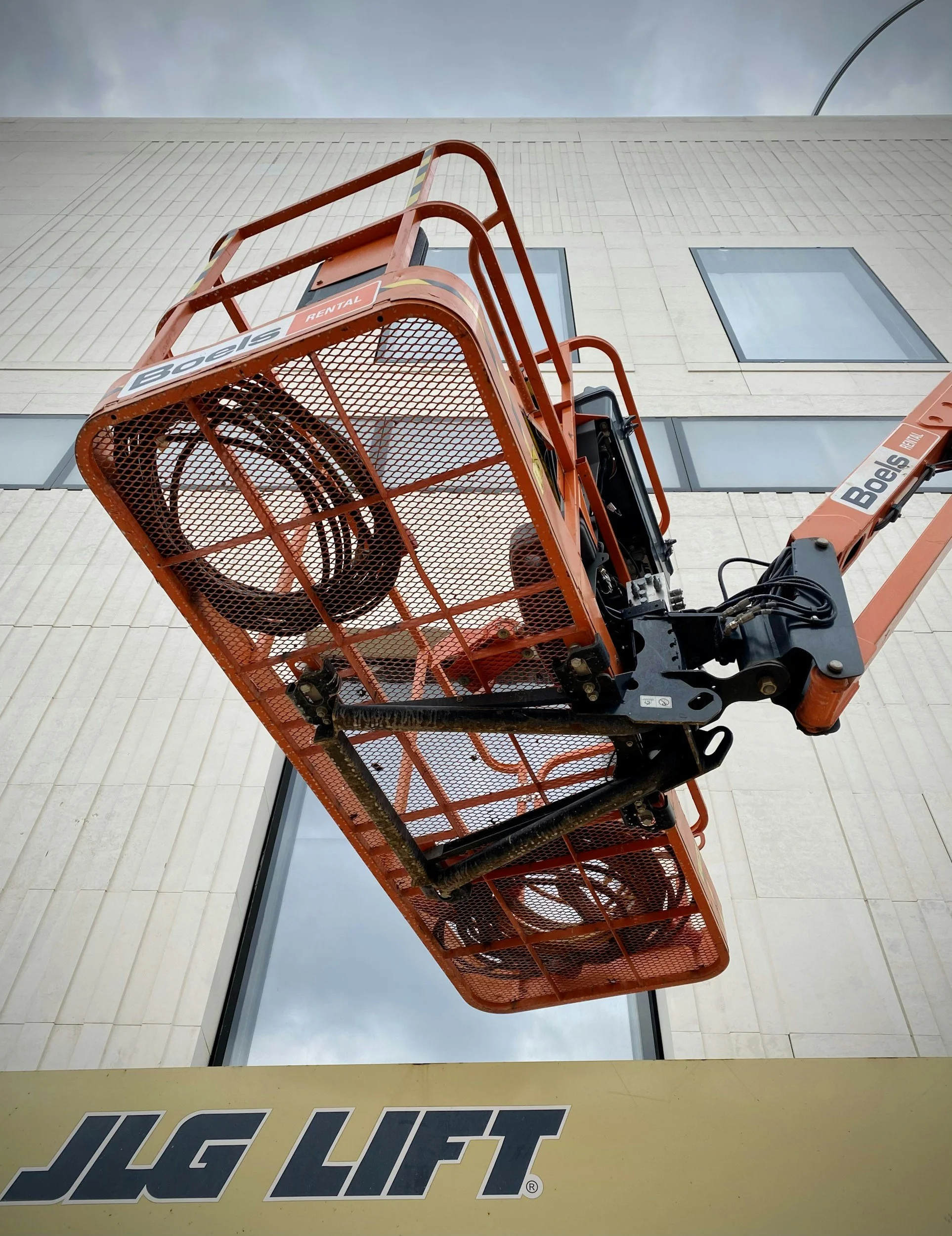 Orange aerial lift platform with black controls, elevated in front of a beige building with rectangular windows, and a cloudy sky in the background.