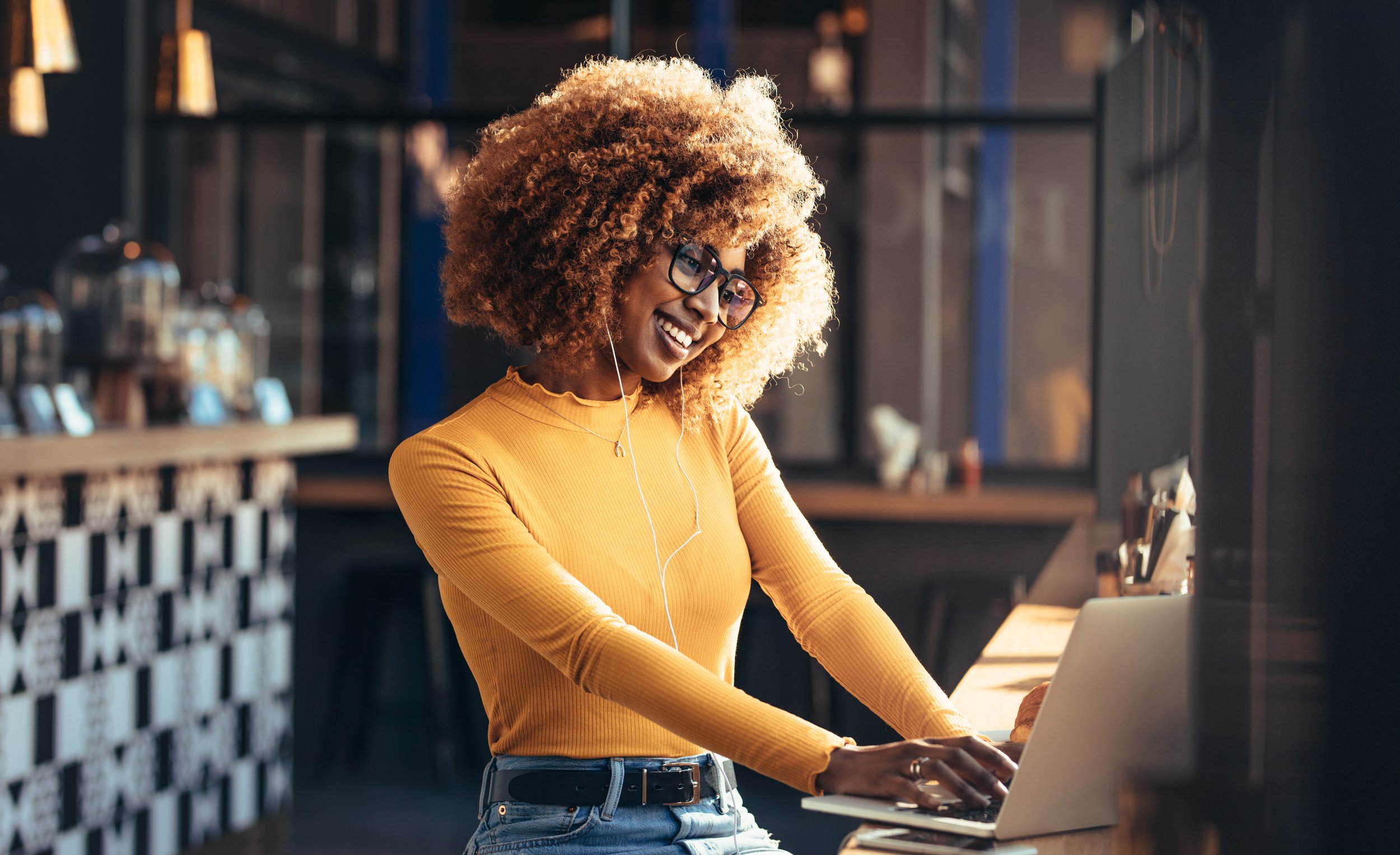 A woman with curly blonde hair, glasses, and a yellow long-sleeve top using a laptop in a modern coffee shop, listening to music with white earbuds.
