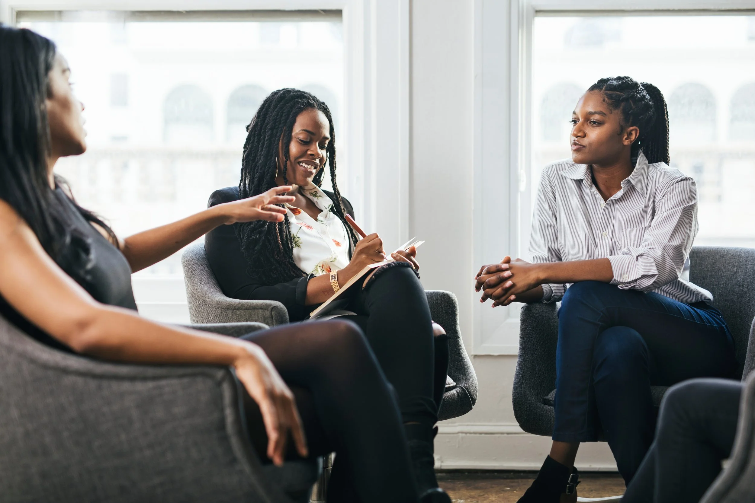 Four women having a discussion in a bright room with large windows, sitting in a semi-circle on gray chairs.