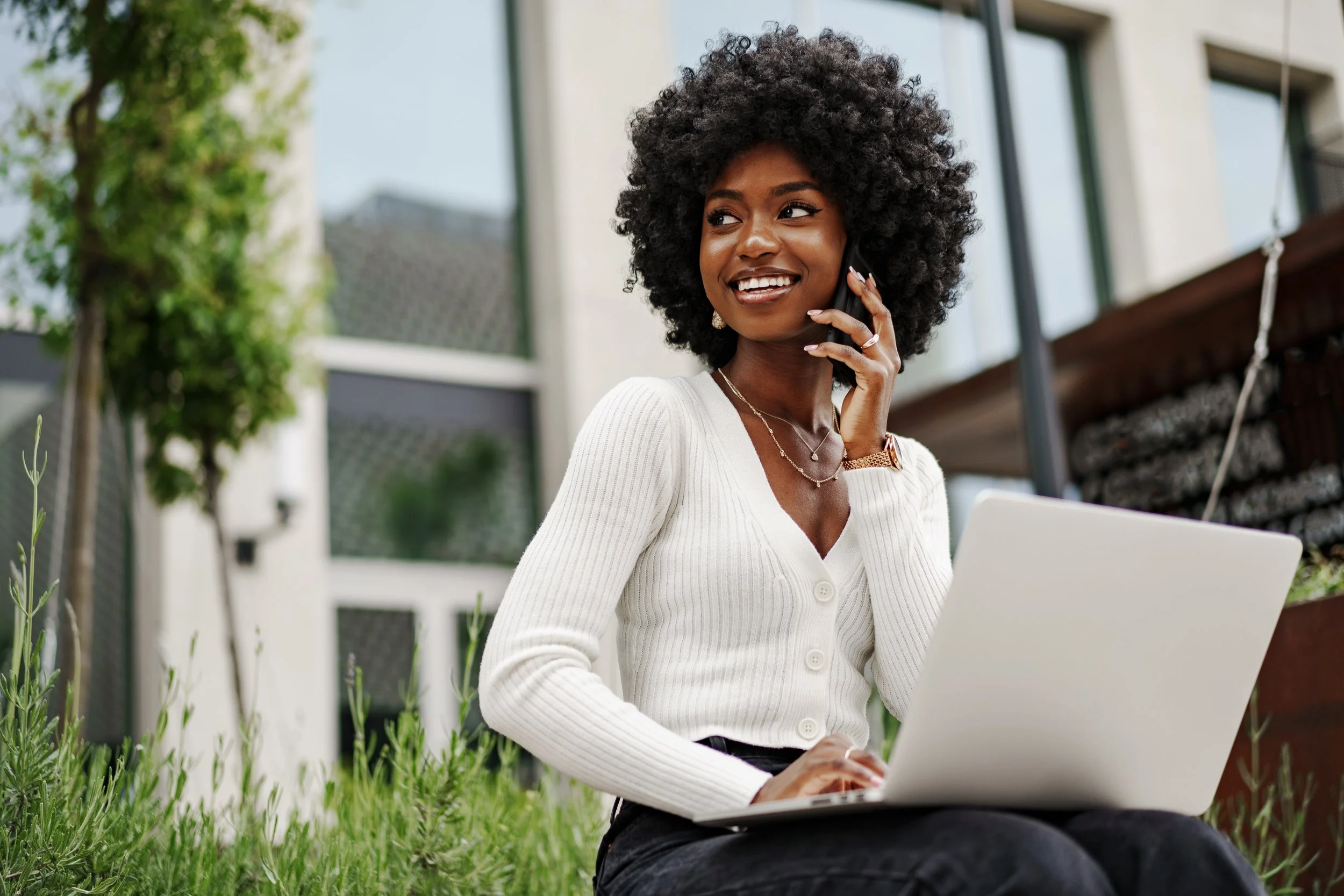 A young woman with curly black hair wearing a white cardigan, sitting outside on grass, using a laptop and talking on her phone.