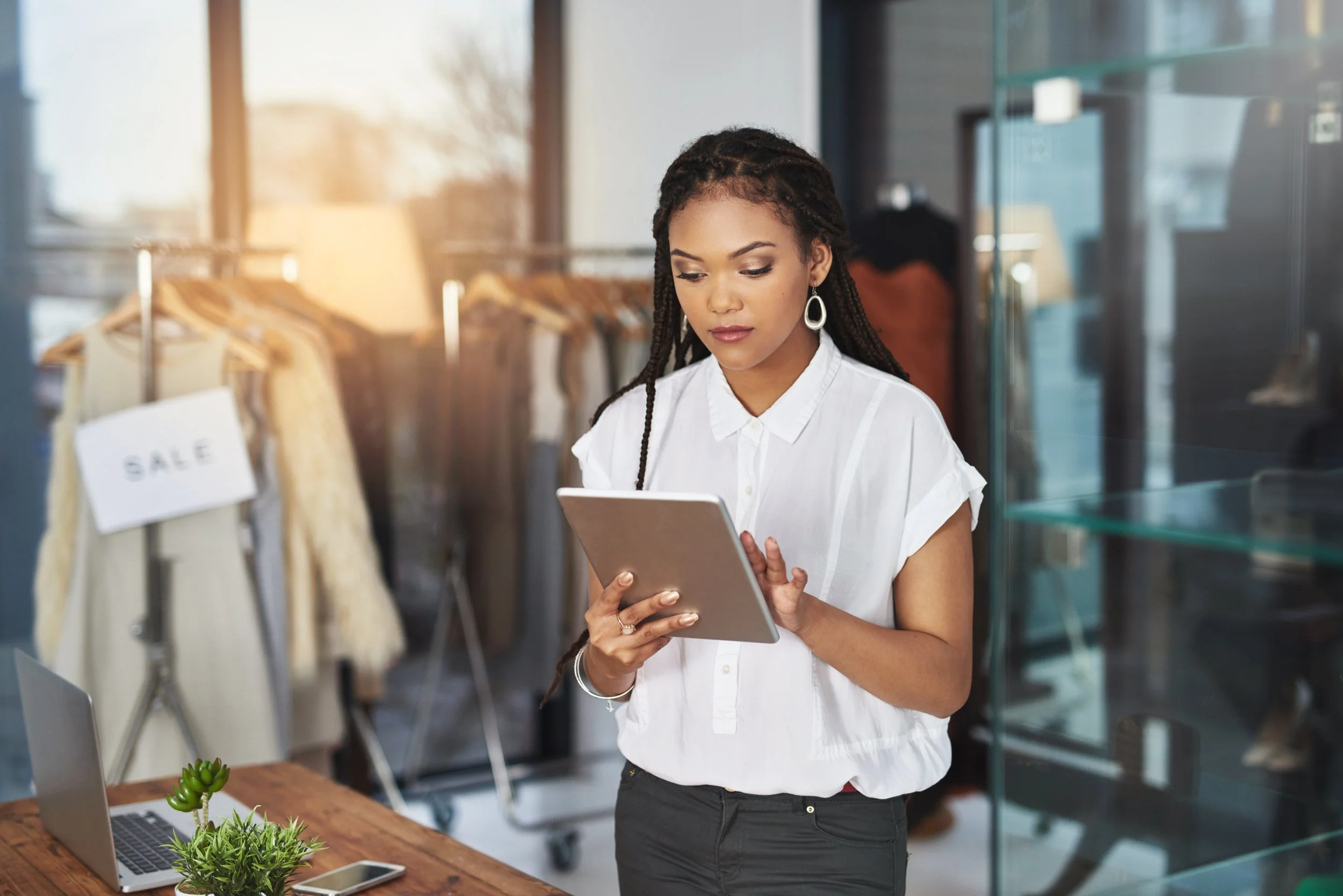 Woman standing in a clothing store, looking at a tablet, with a laptop, smartphone, and plant on a wooden table nearby, and clothing racks with sale signs in the background.