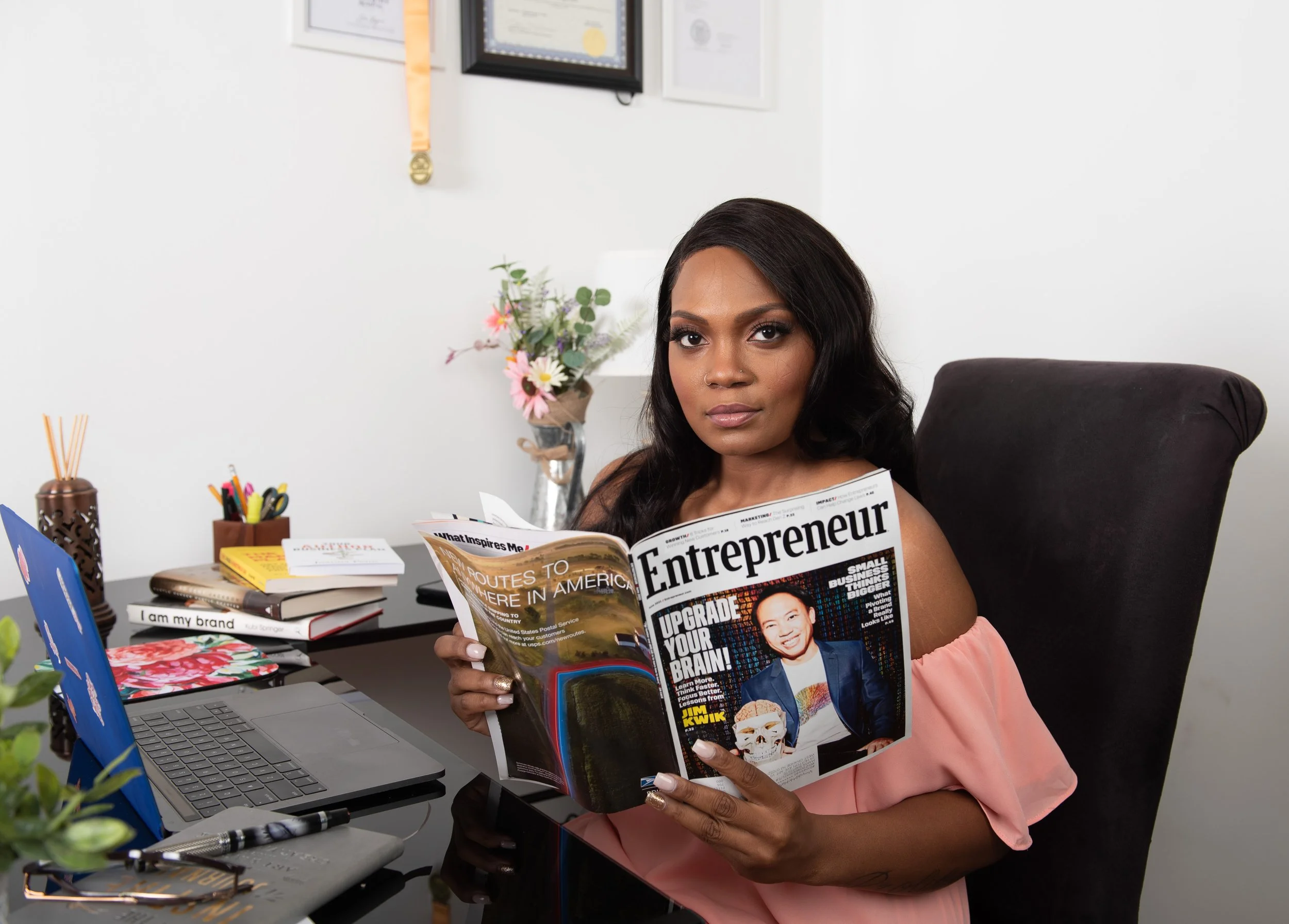 A woman sitting at a desk holding a magazine titled 'Entrepreneur' with a headline 'Upgrade Your Brain!'. She is in an office with framed certificates, a flower vase, and office supplies on the desk.