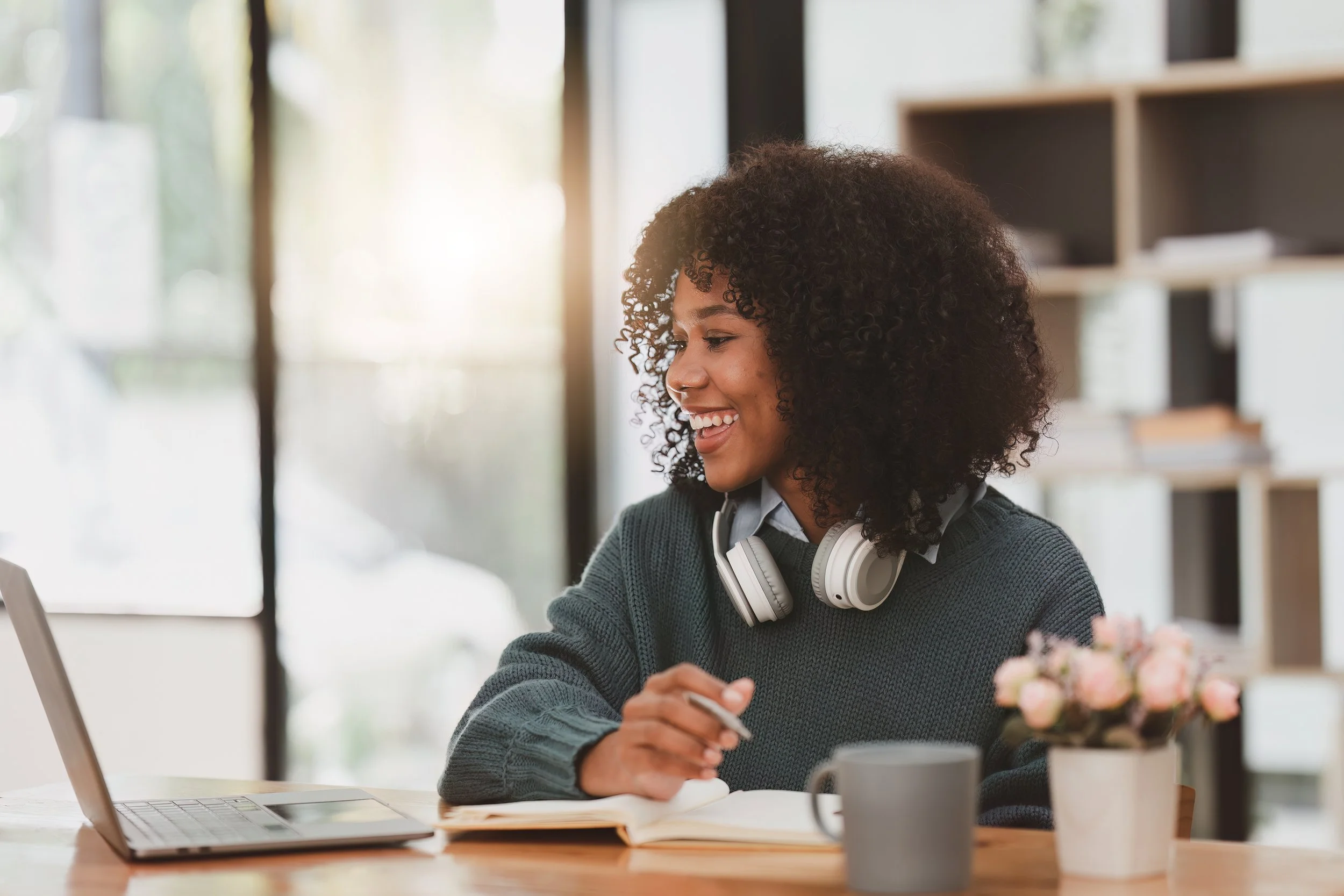 A young woman with curly hair and headphones around her neck smiling while working at a desk with a laptop, a notebook, a gray mug, and a small pink flower arrangement in a vase inside a well-lit room.