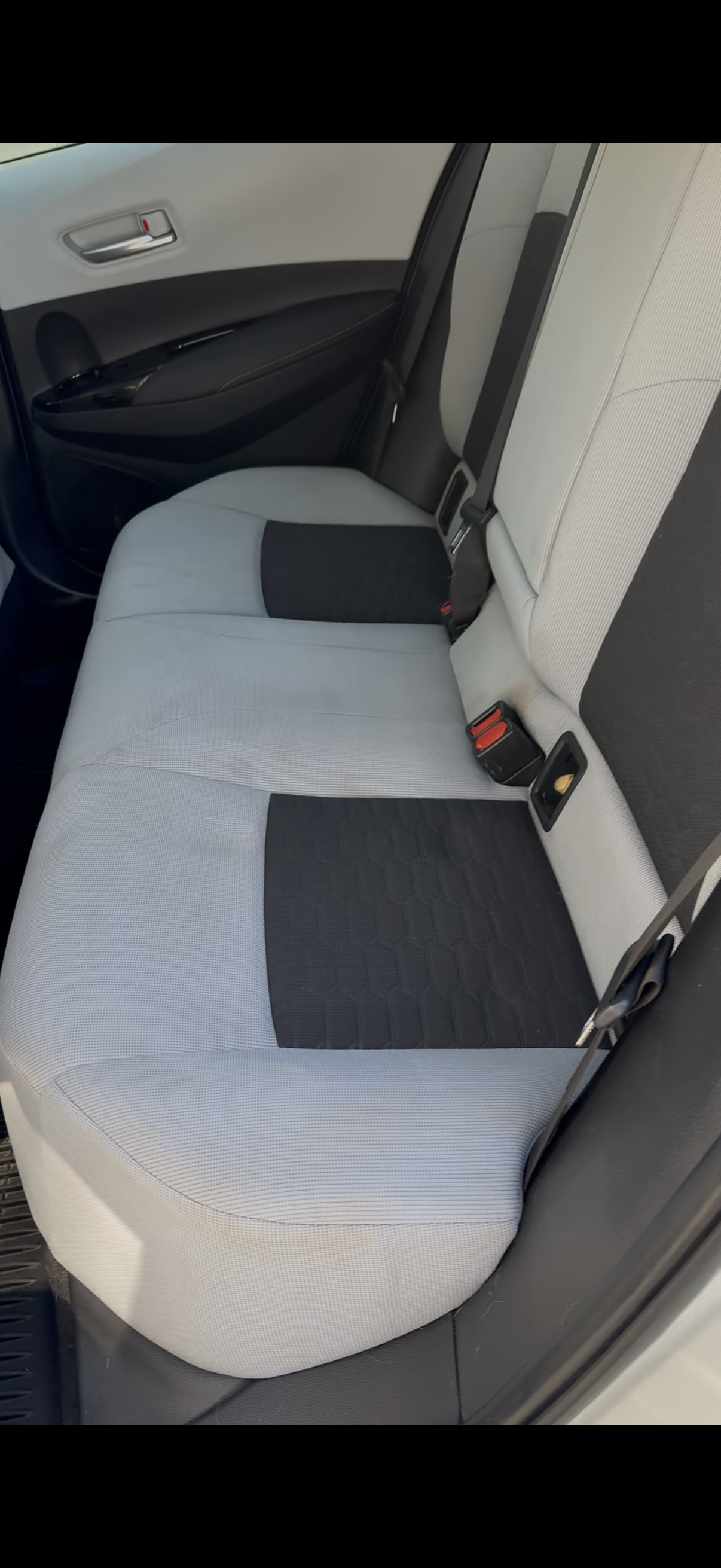 The back seat of a car with light-colored upholstery, black seat inserts, three seat belts, and a window on the door.