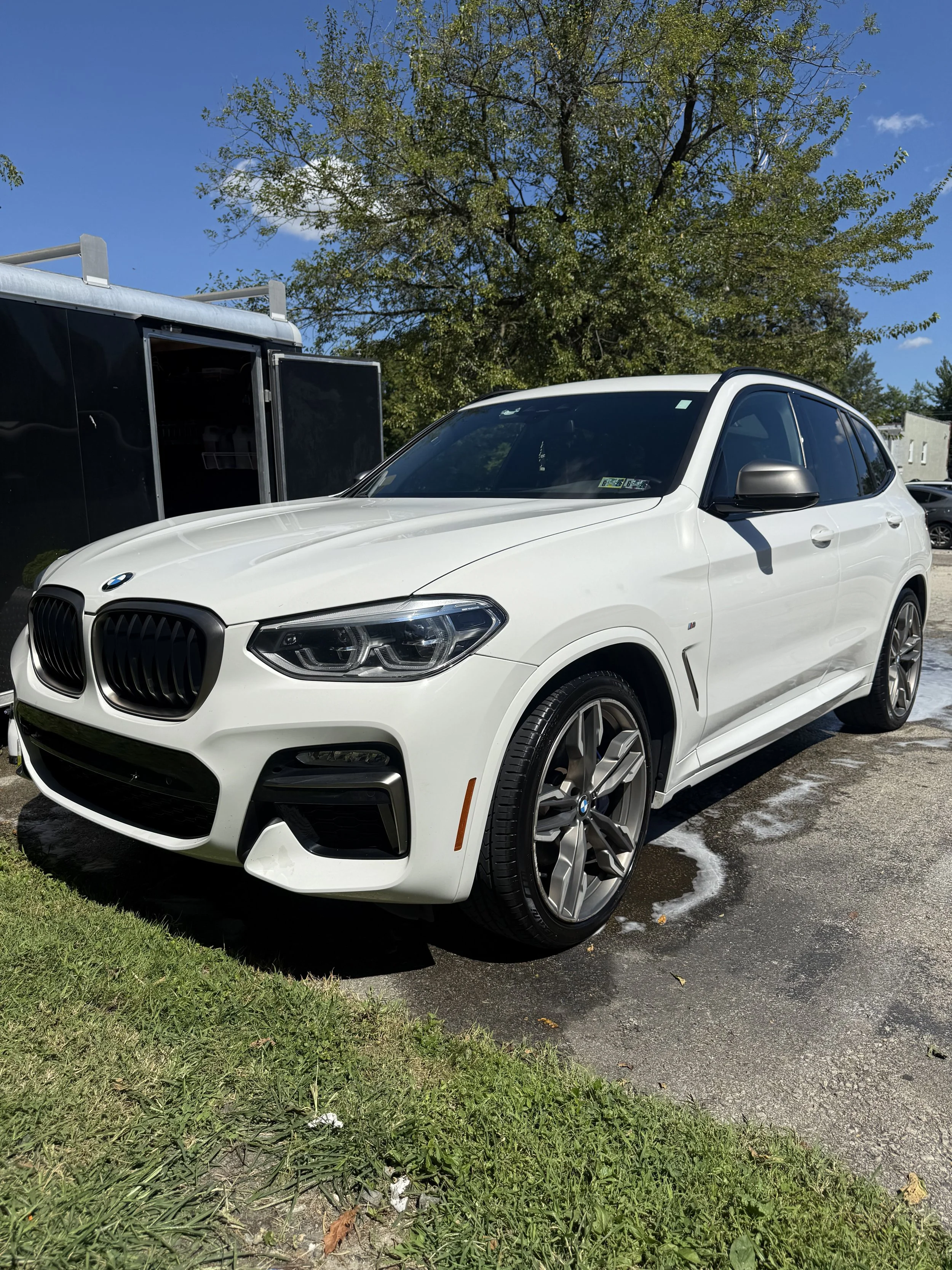 A white BMW SUV parked on a wet asphalt surface with soap suds around the tires, next to a grassy area and a large tree in the background on a sunny day.