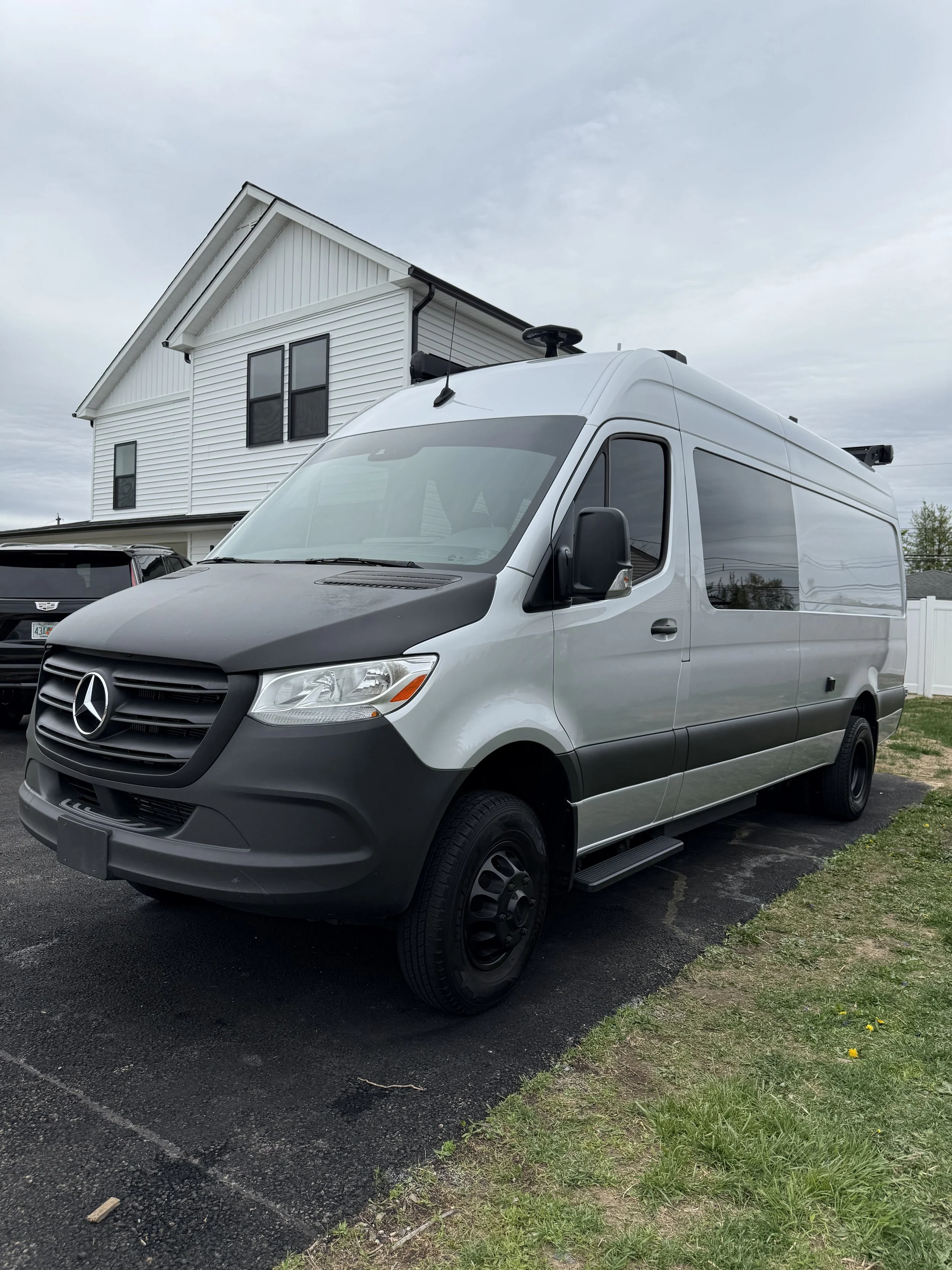 Silver Mercedes-Benz Sprinter van parked on a driveway in front of a white house on an overcast day.