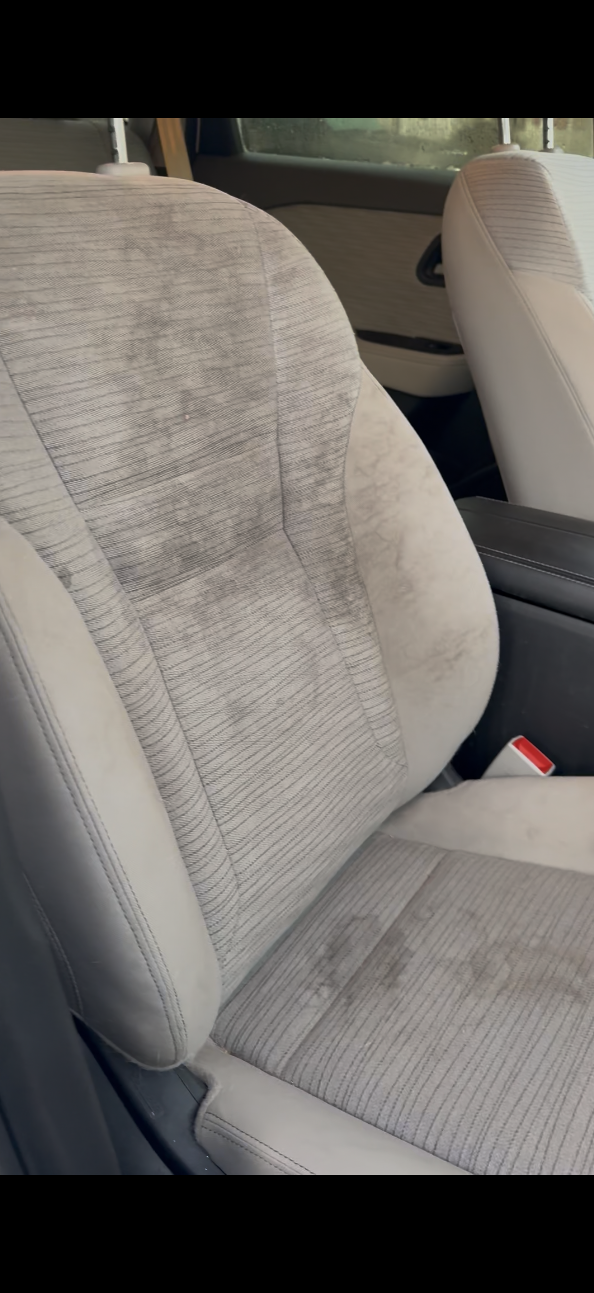 The image shows the front and back seats of a car with light-colored fabric upholstery, some dirt or stains visible on the seats, and a red seatbelt buckle.