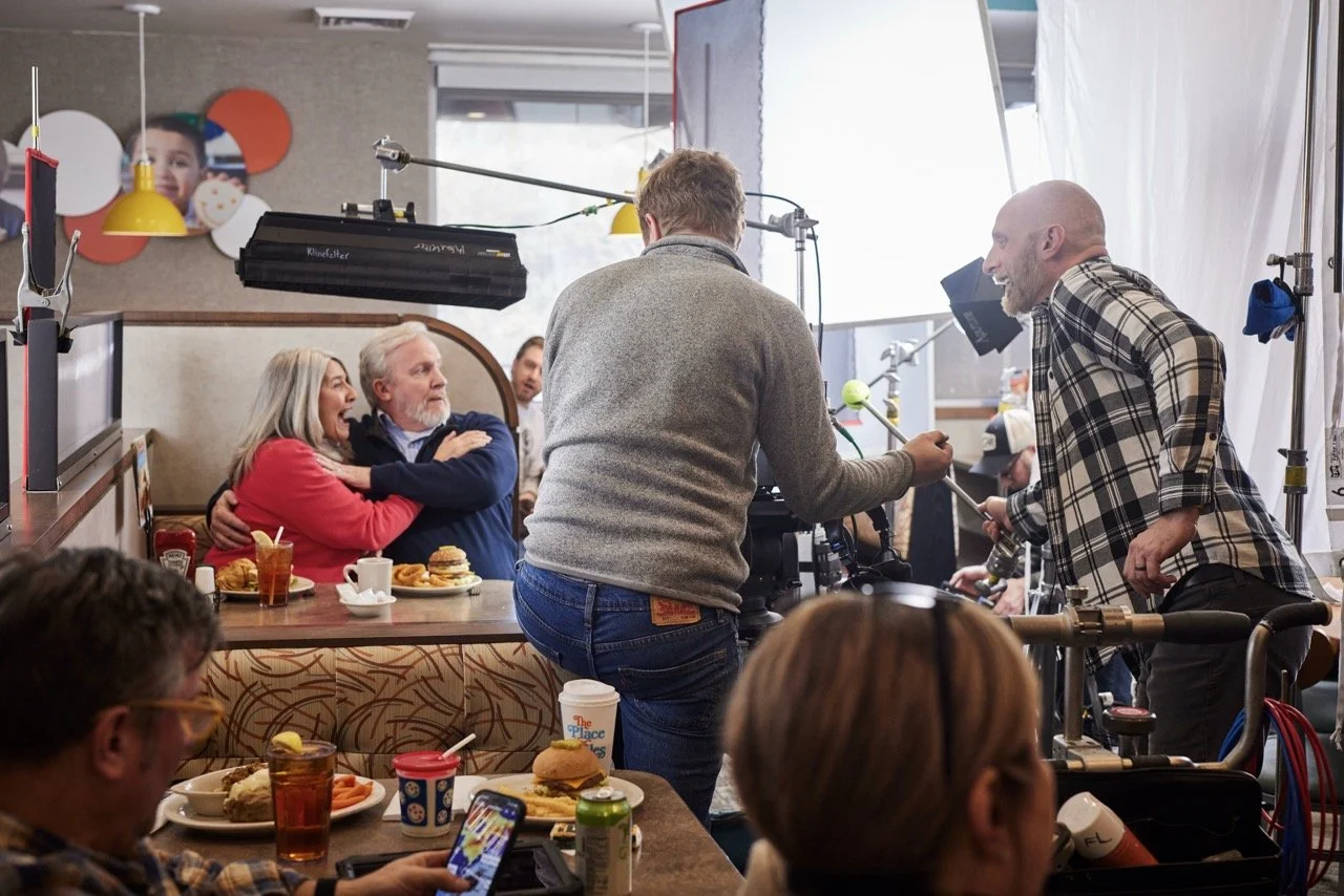Video production film set, with a actors sitting at a restaurant table, while a crew member operates equipment nearby.