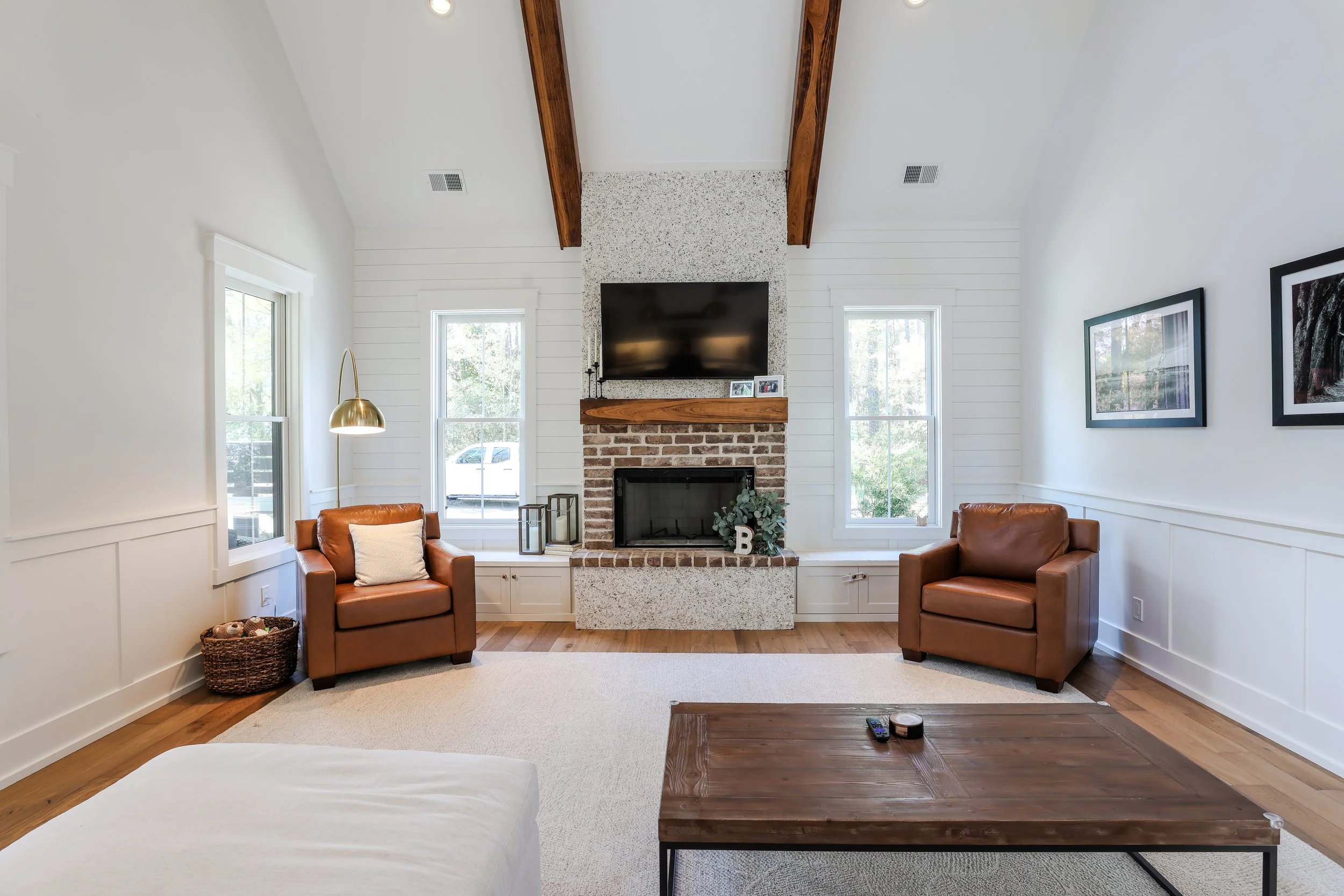 Living room with white walls, exposed wooden beams, a brick and concrete fireplace with a TV above, two brown leather armchairs, a wooden coffee table, and framed artwork on the wall.