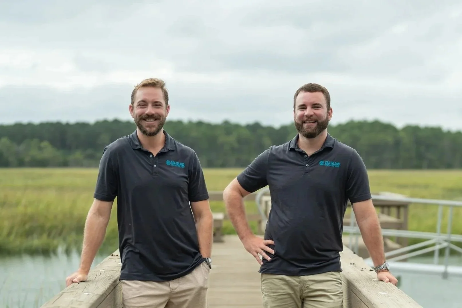 Two smiling men standing on a wooden dock outdoors, wearing dark polo shirts with a small logo that reads 'Sea Island'. They have beards and are in a lush green landscape with water and trees in the background.