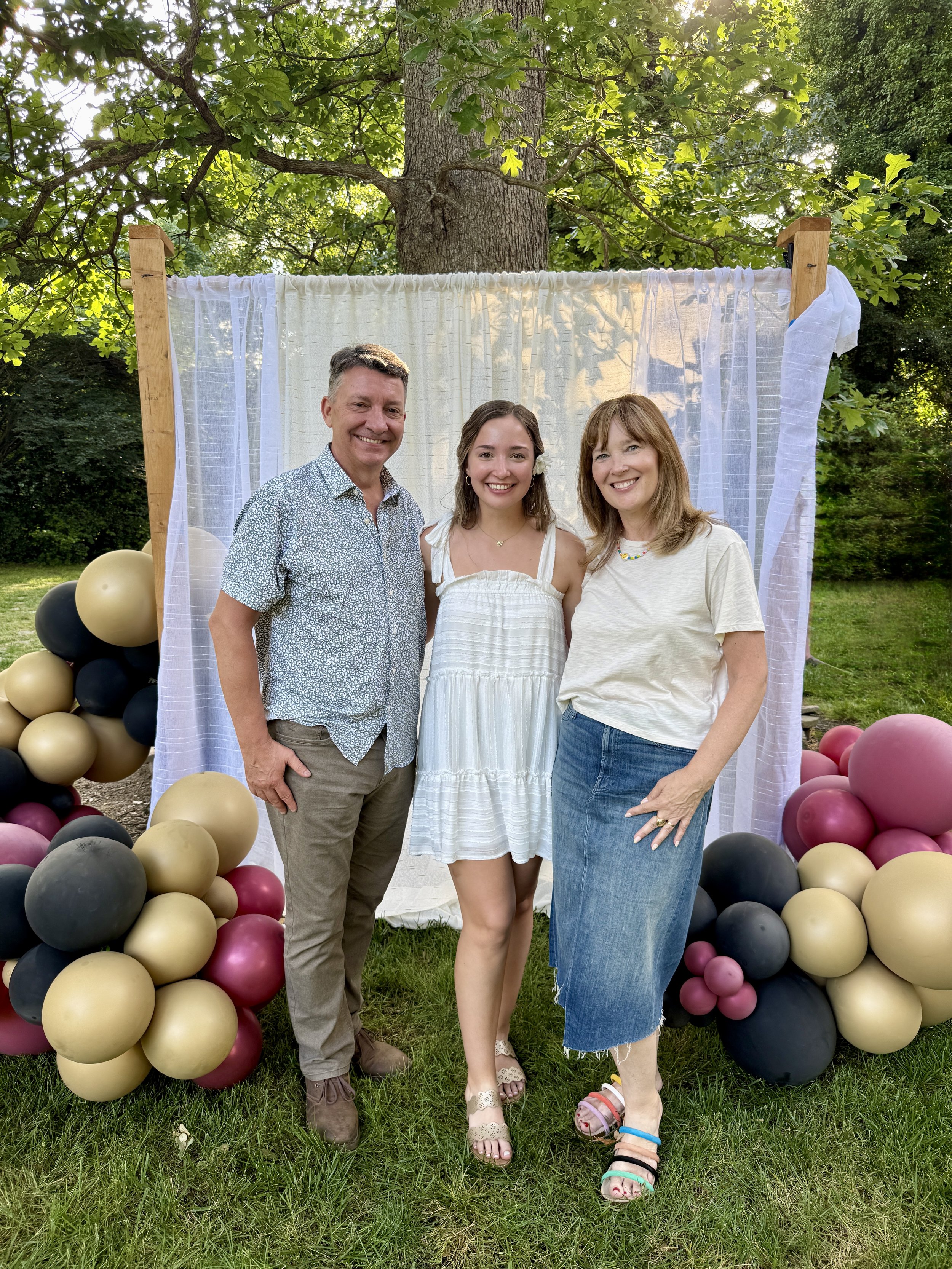 Three smiling people standing outdoors in front of a decorated backdrop with balloons on each side.