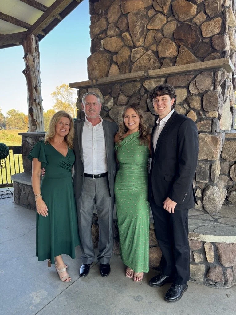 Four people dressed in formal attire standing together in front of a large stone fireplace on a covered porch, with a scenic outdoor background.