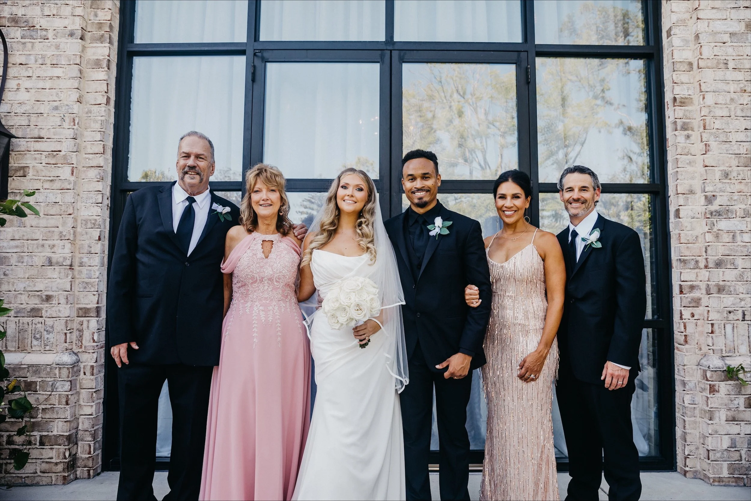 Group of six people in formal attire standing outside a building with brick walls and large black-framed windows, smiling for a photo.