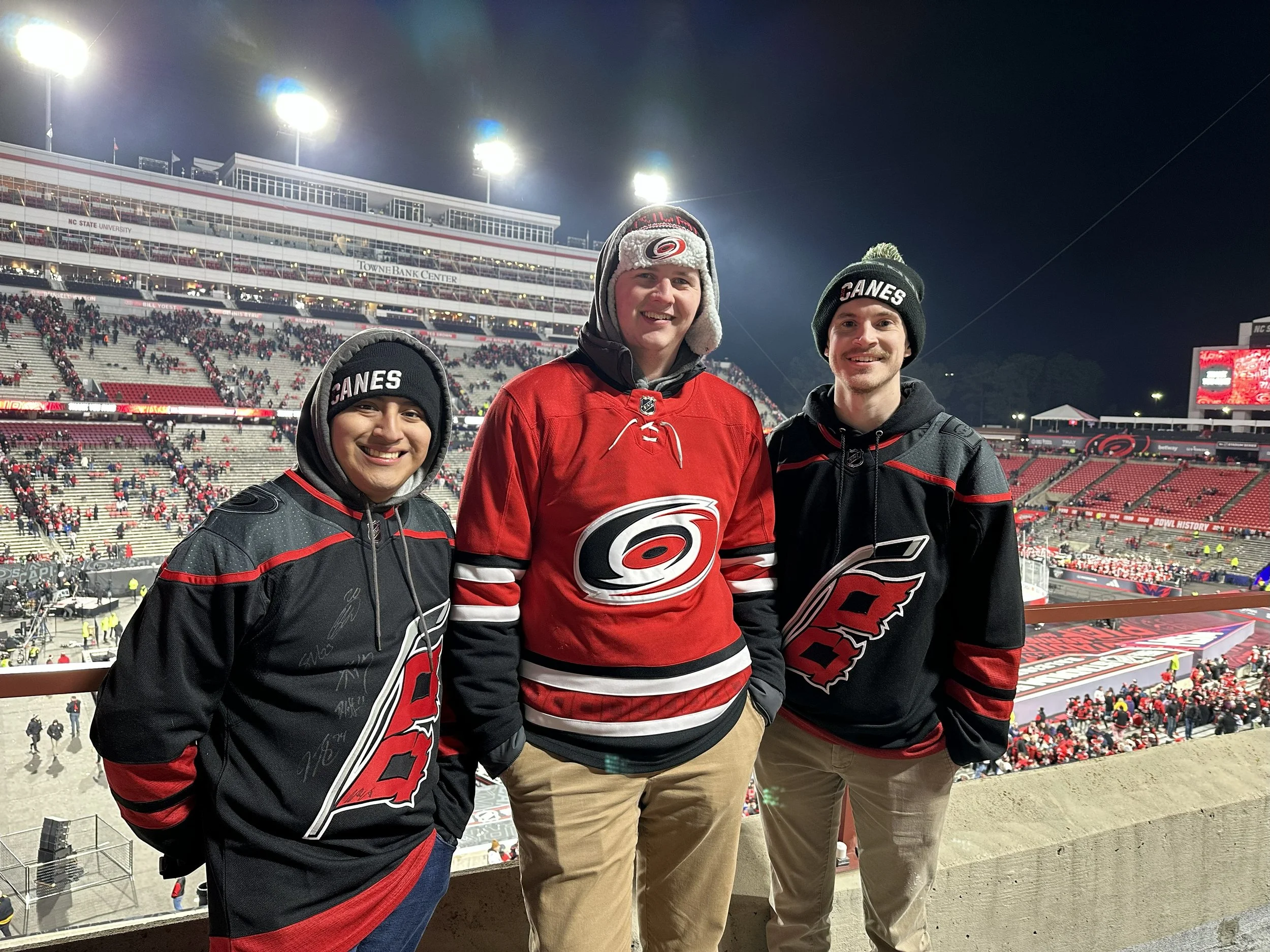 Three young men at a stadium during a hockey game, wearing Carolina Hurricanes hockey jerseys and hats, standing near the railing with fans and the arena filled with spectators in the background.
