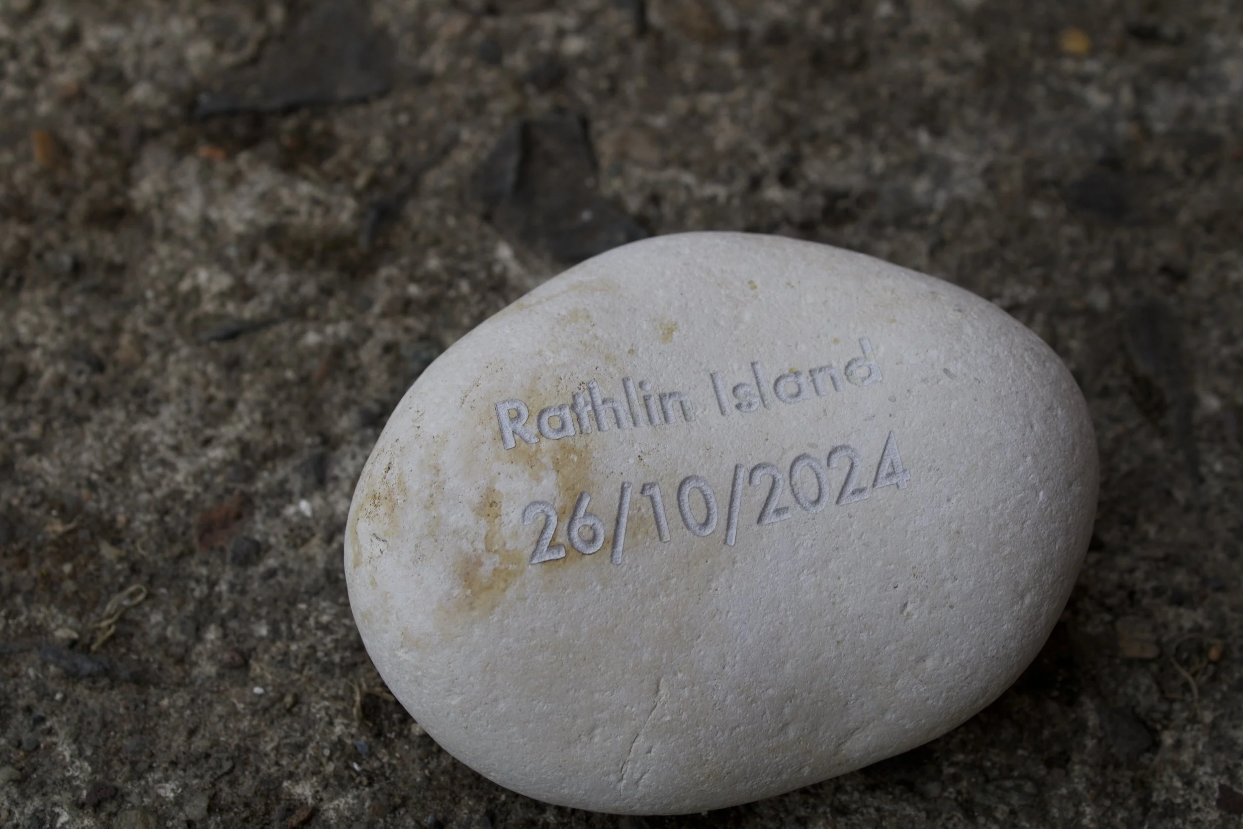 A white stone with engraved text reading 'Rathlin Island 26/10/2024' on a sandy surface.