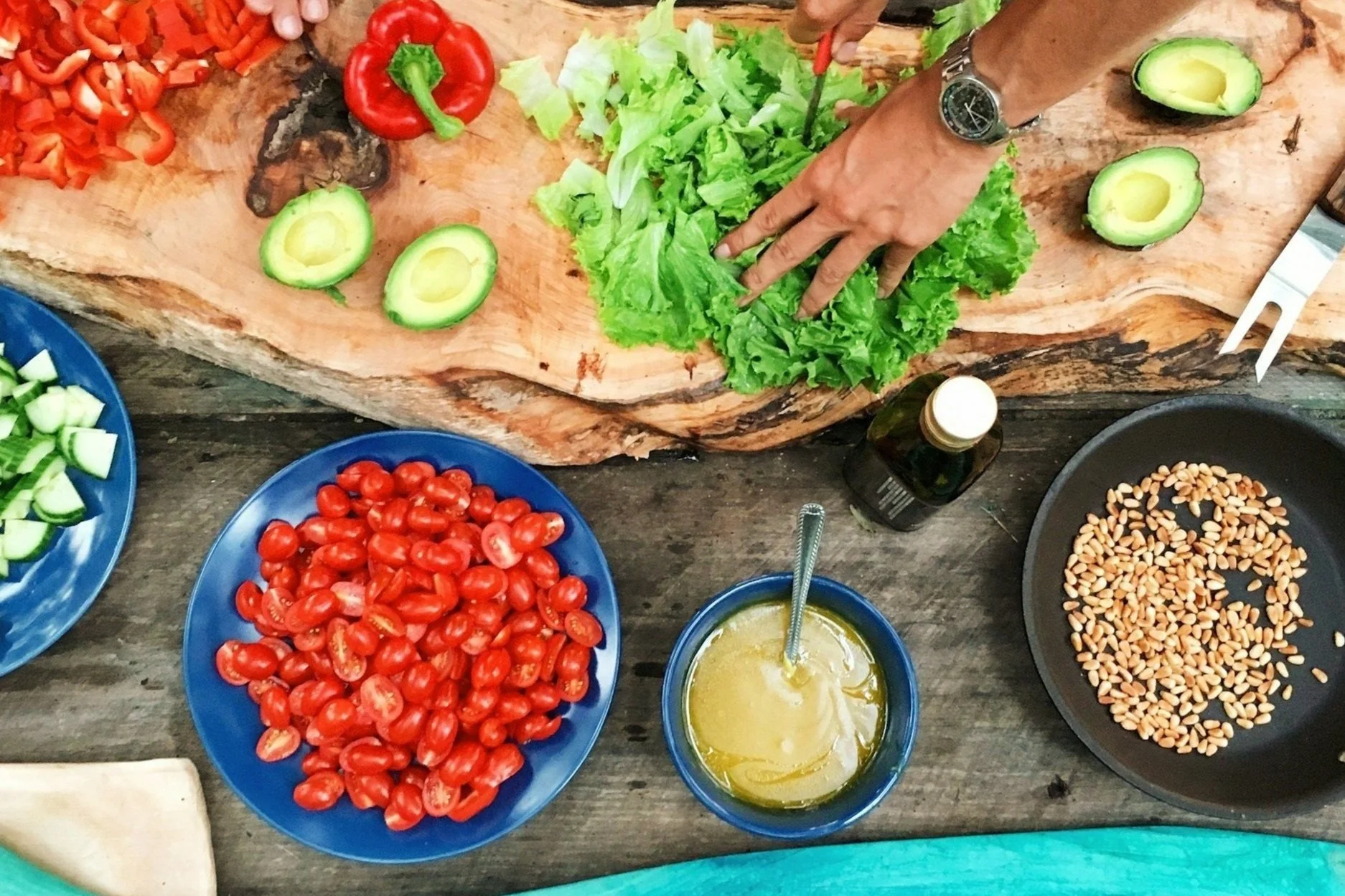 A person chopping lettuce on a wooden cutting board with sliced avocado, cherry tomatoes, chopped red bell peppers, cucumbers, pine nuts, and a bowl of dressing nearby.