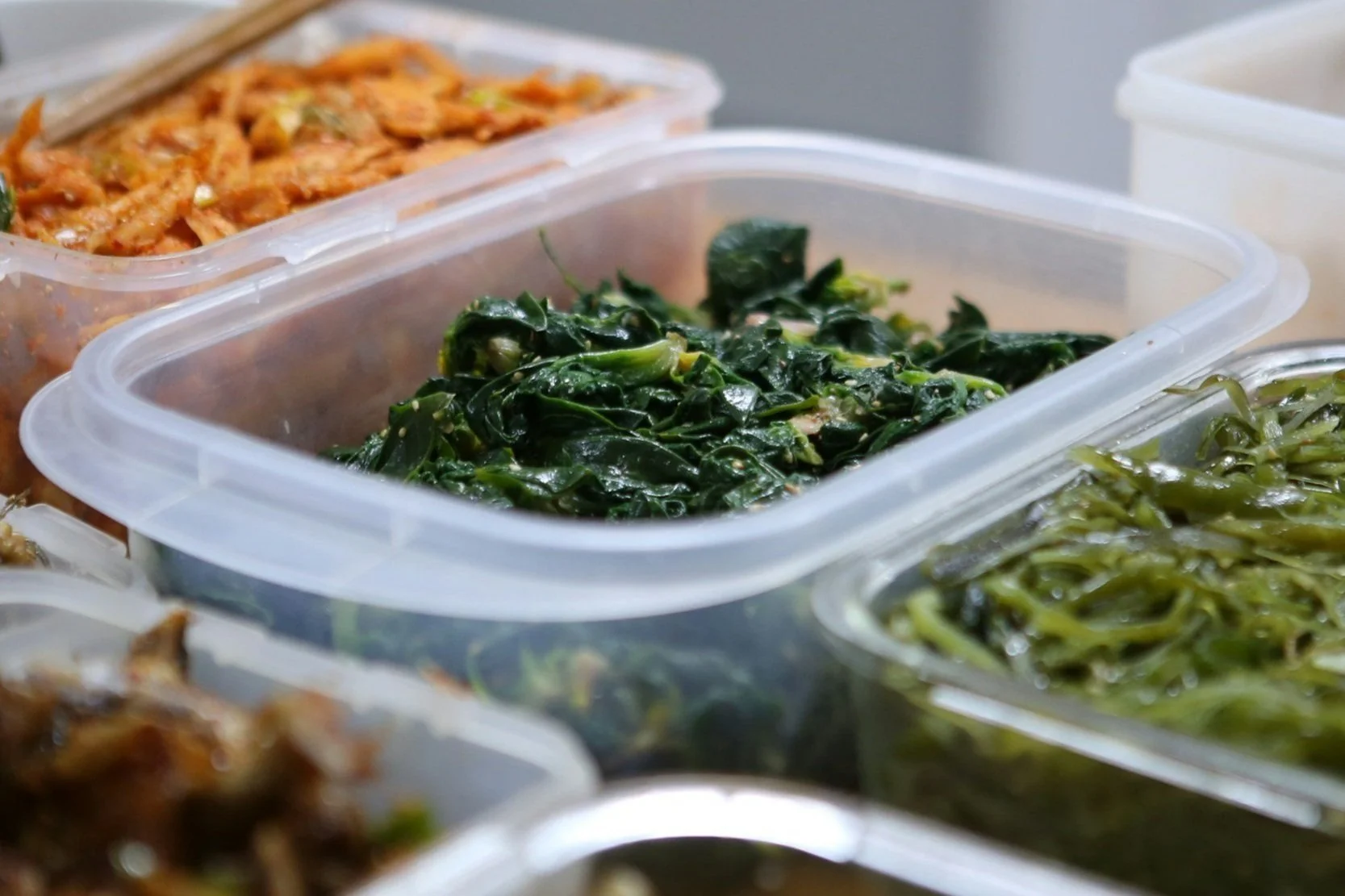 Plastic containers filled with Korean side dishes, including seasoned spinach, kimchi, and marinated vegetables.