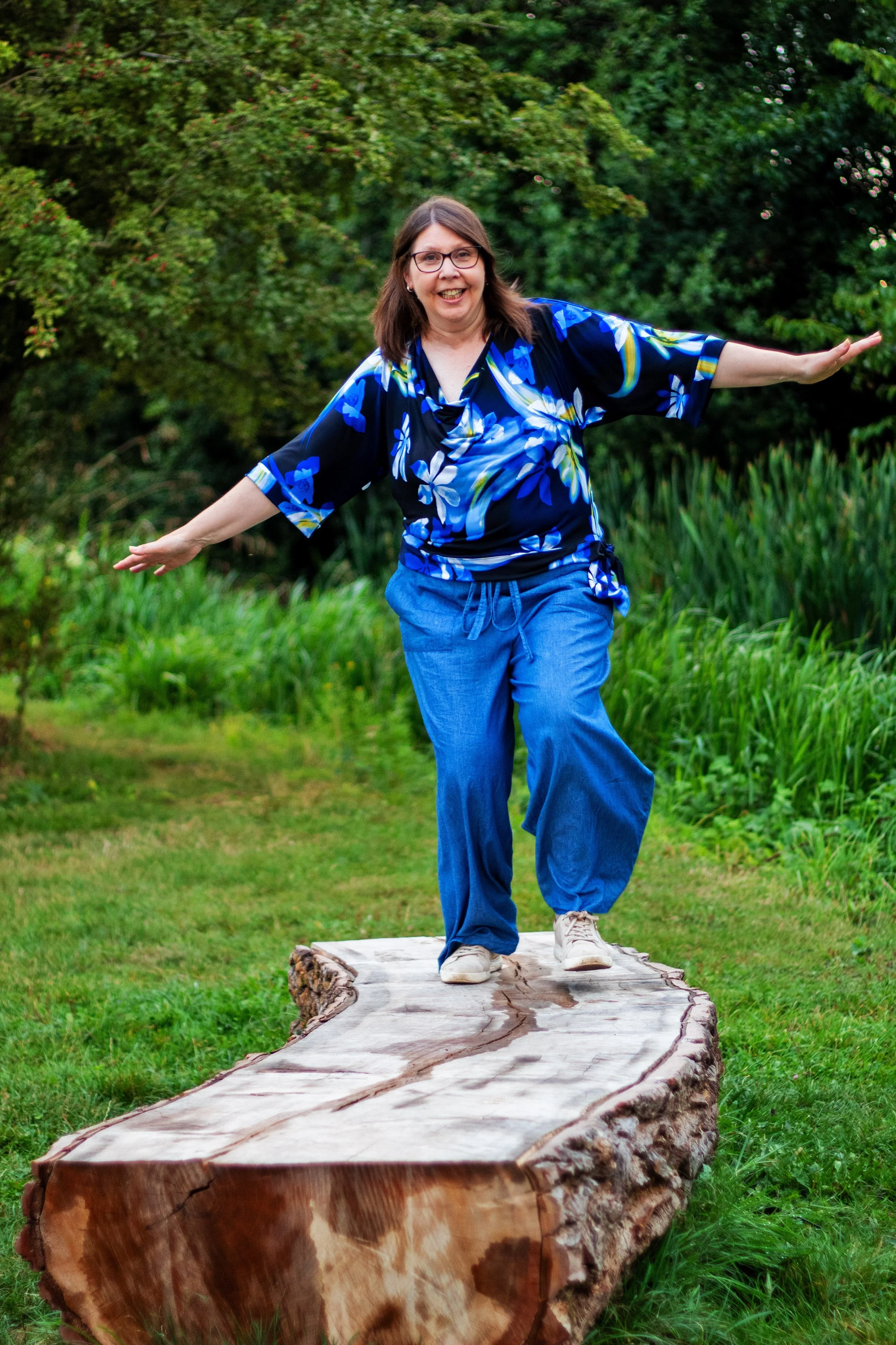 Woman with glasses and a blue floral top balancing on a large wooden log in a lush green outdoor setting.