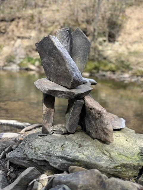 Stacked rocks balance on a large flat rock by a creek with a wooded background.