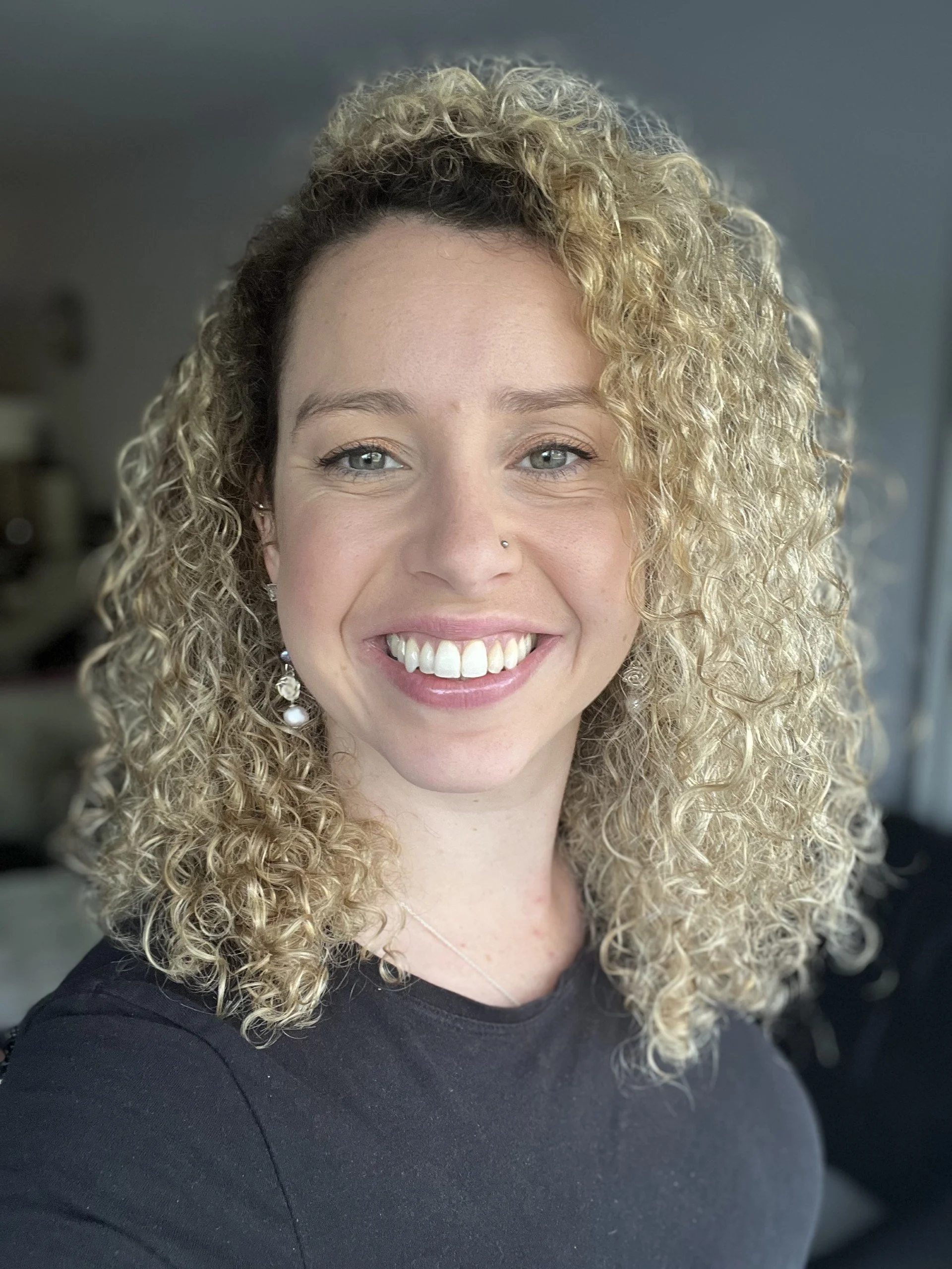 A woman with curly blonde hair, light skin, wearing a black top, smiling, with light makeup, earrings, and a nose piercing, indoors.