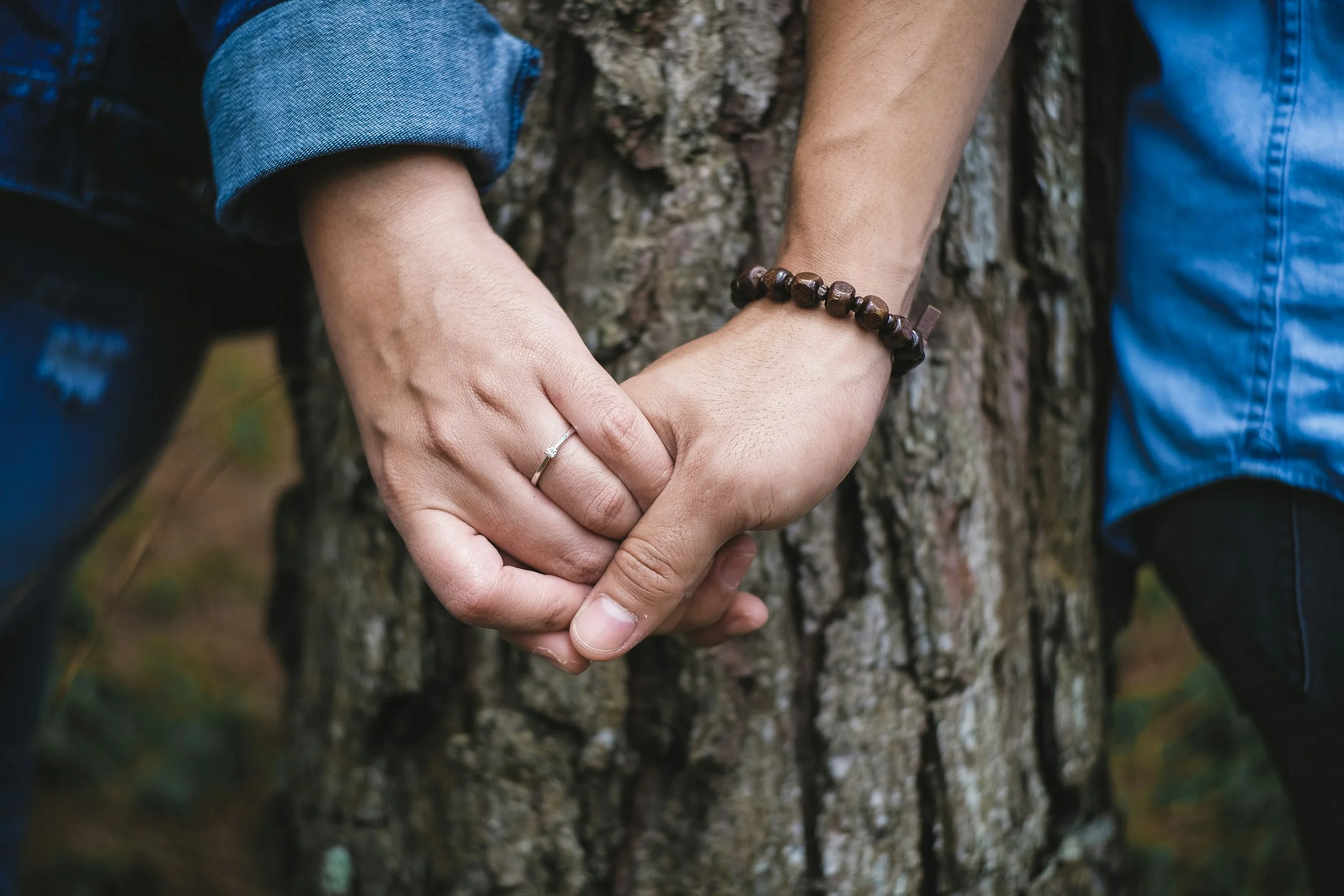 Two people holding hands against a tree trunk, one person wearing a ring and a beaded bracelet.