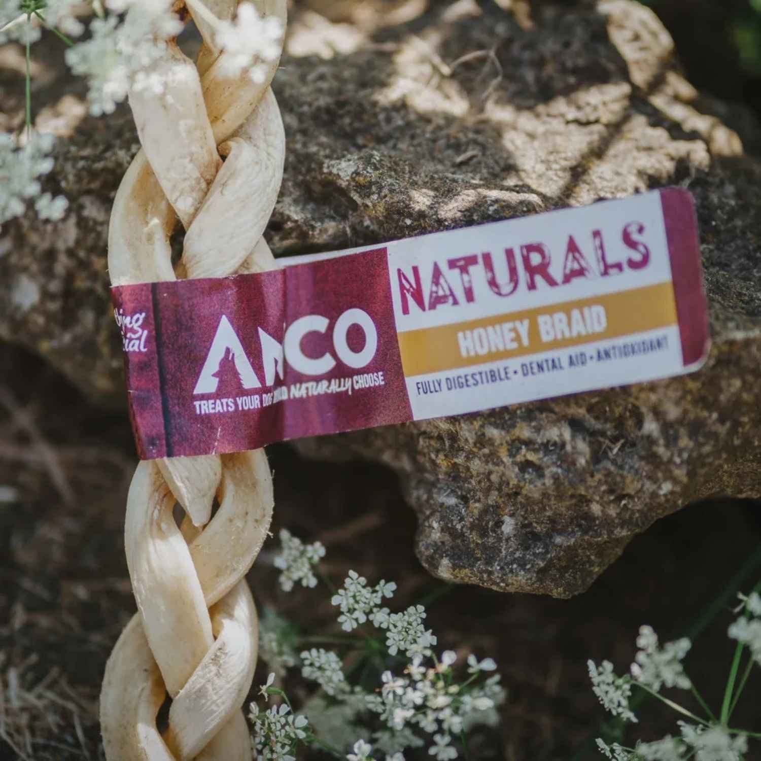 Close-up of an Anco Naturals Honey Braid chew stick with its branded maroon and gold label band, resting outdoors near rocks and small white flowers.