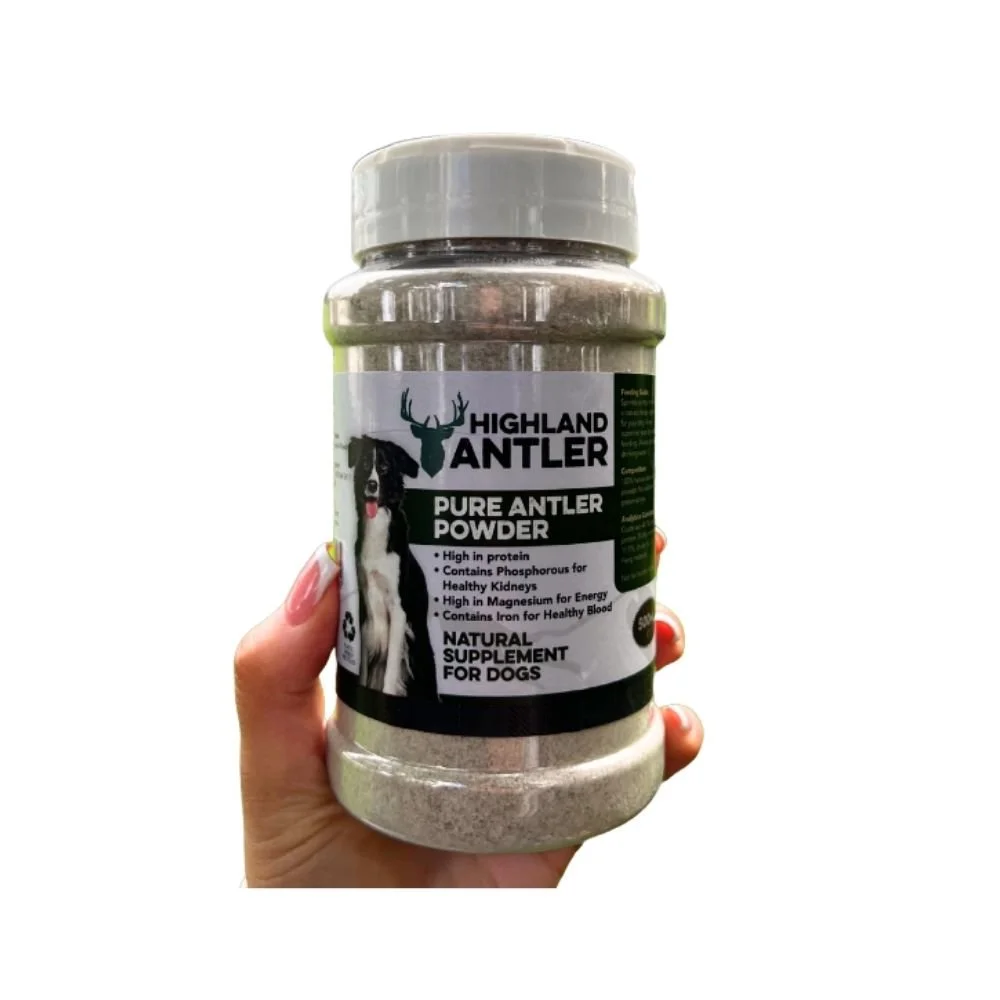 A person holding a 500ml shaker bottle of Highland Antler Pure Antler Powder natural dog supplement against a white background.
