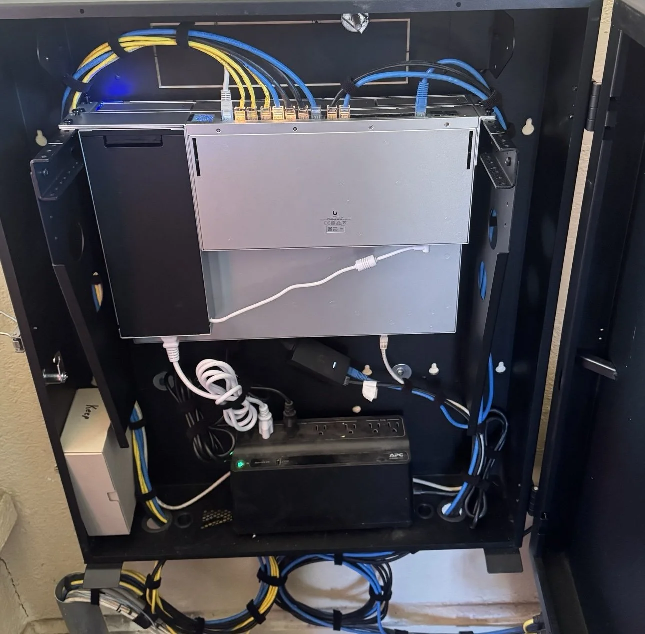 Interior of a black network or electronic equipment cabinet showing power and data cables connected to a silver server or switch, with additional components and wiring visible inside.