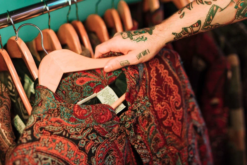 Person with tattoos on arm holding a paisley-patterned garment on a hanger in a clothing store.