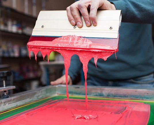Person holding a squeegee with pink screen printing ink, preparing to print on a T-shirt.