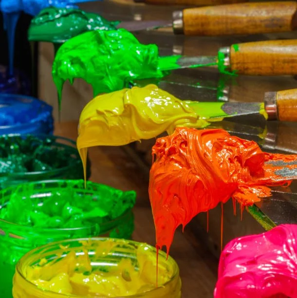 Painted green, yellow, orange, and pink oil paints on brushes with open jars of matching paints in the background.