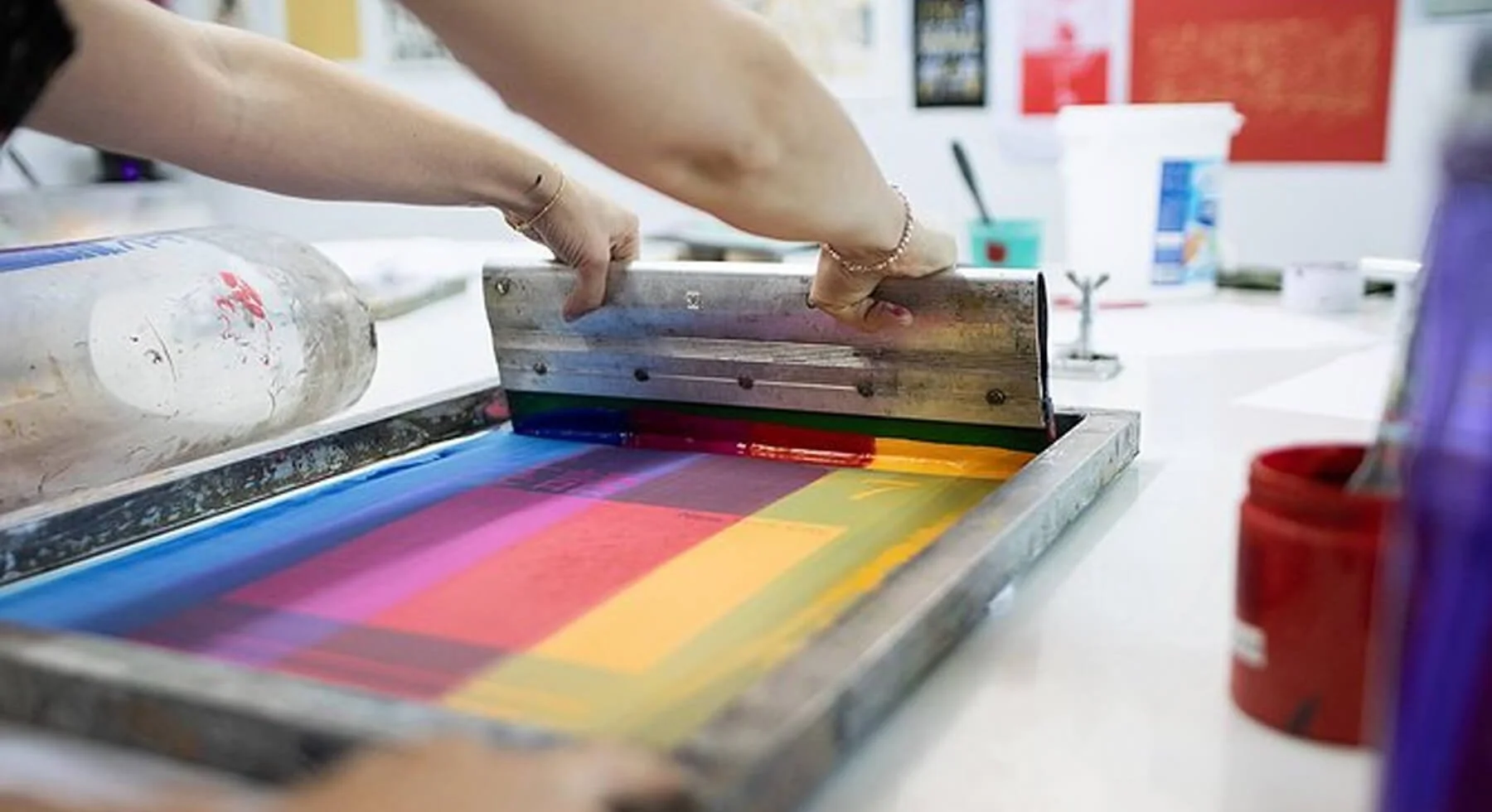 Person using a printmaking or screen-printing squeegee on a colorful inked screen for printing.