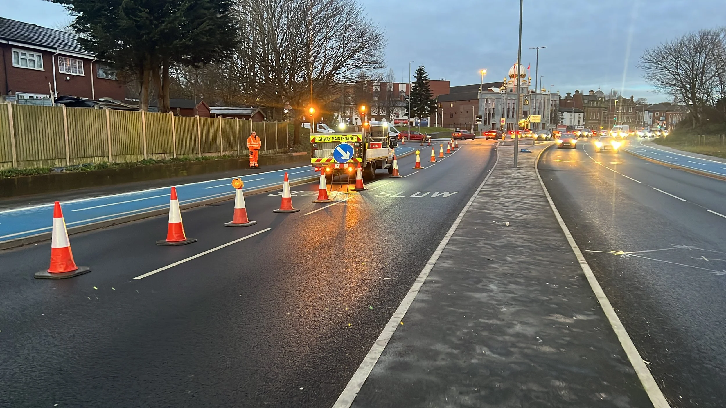 A road under maintenance with traffic cones and a work vehicle, a worker in orange safety gear, and traffic on a wet road in a city setting during dusk.