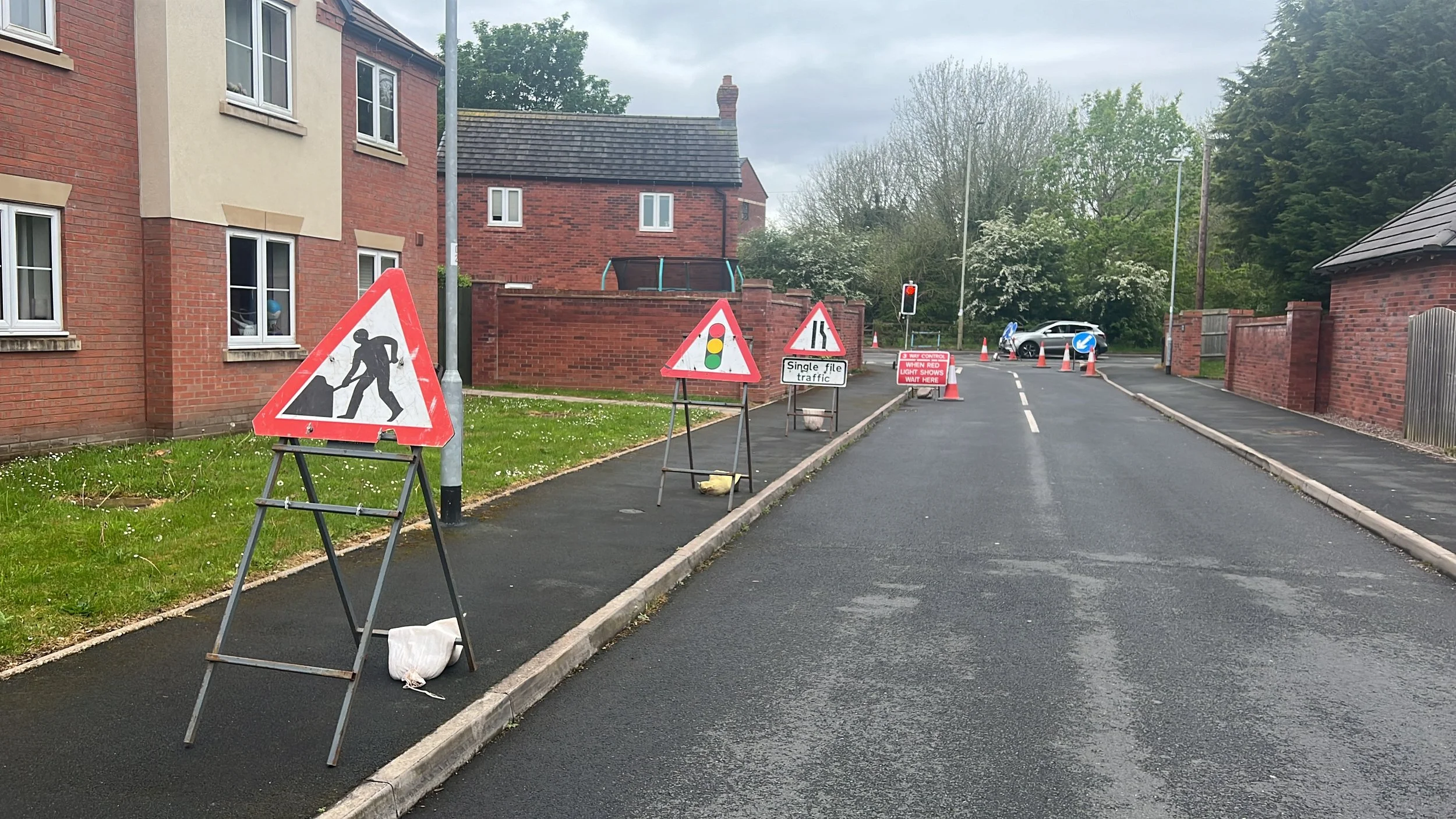 Construction signs and traffic cones blocking a residential street under construction, with a traffic light and parked cars in the background.
