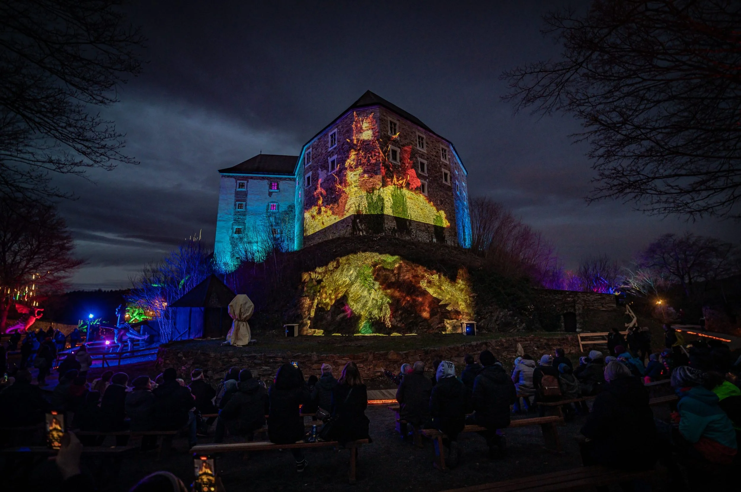Nighttime crowd watching a castle illuminated with colorful projection mapping.