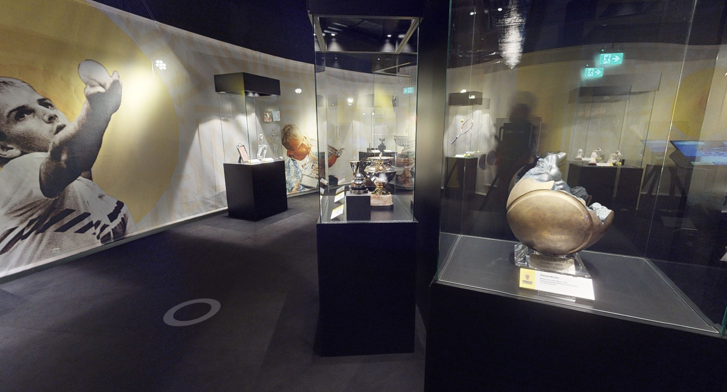 Museum exhibit with vintage trophies and artifacts inside display cases, large artwork of a football player on the wall.