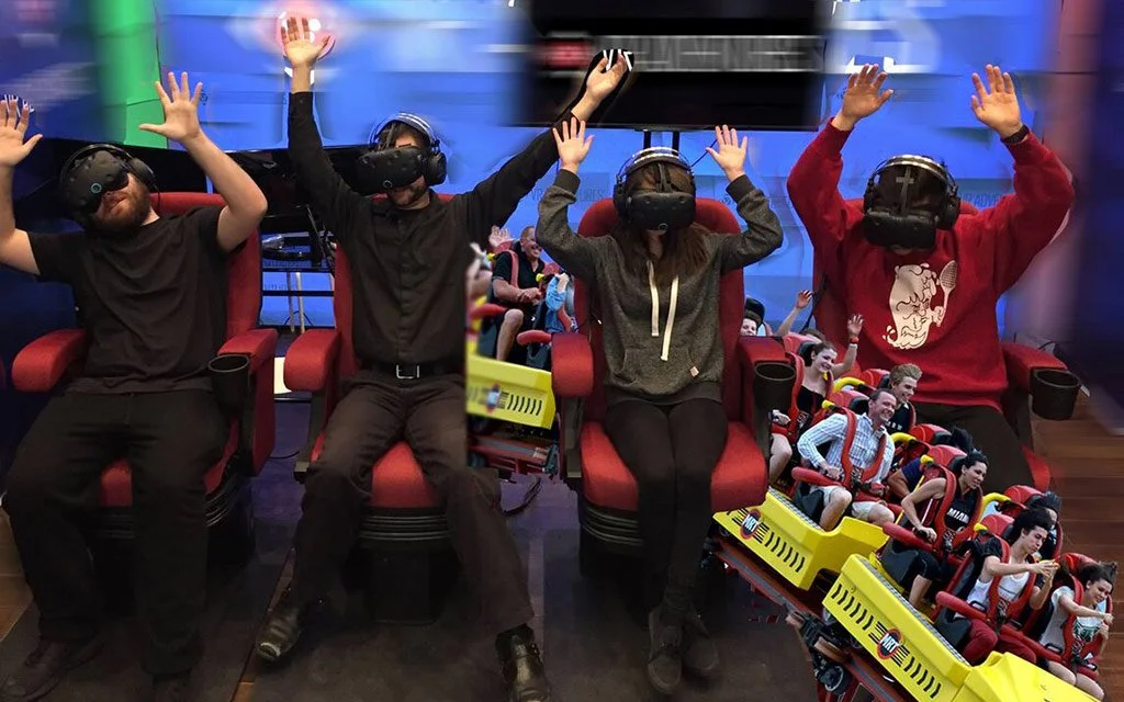 Four people seated in a row wearing virtual reality headsets, with their hands raised, attached to a virtual roller coaster ride simulation; on their right, people on a physical roller coaster ride are visible, creating a themed amusement park atmosp