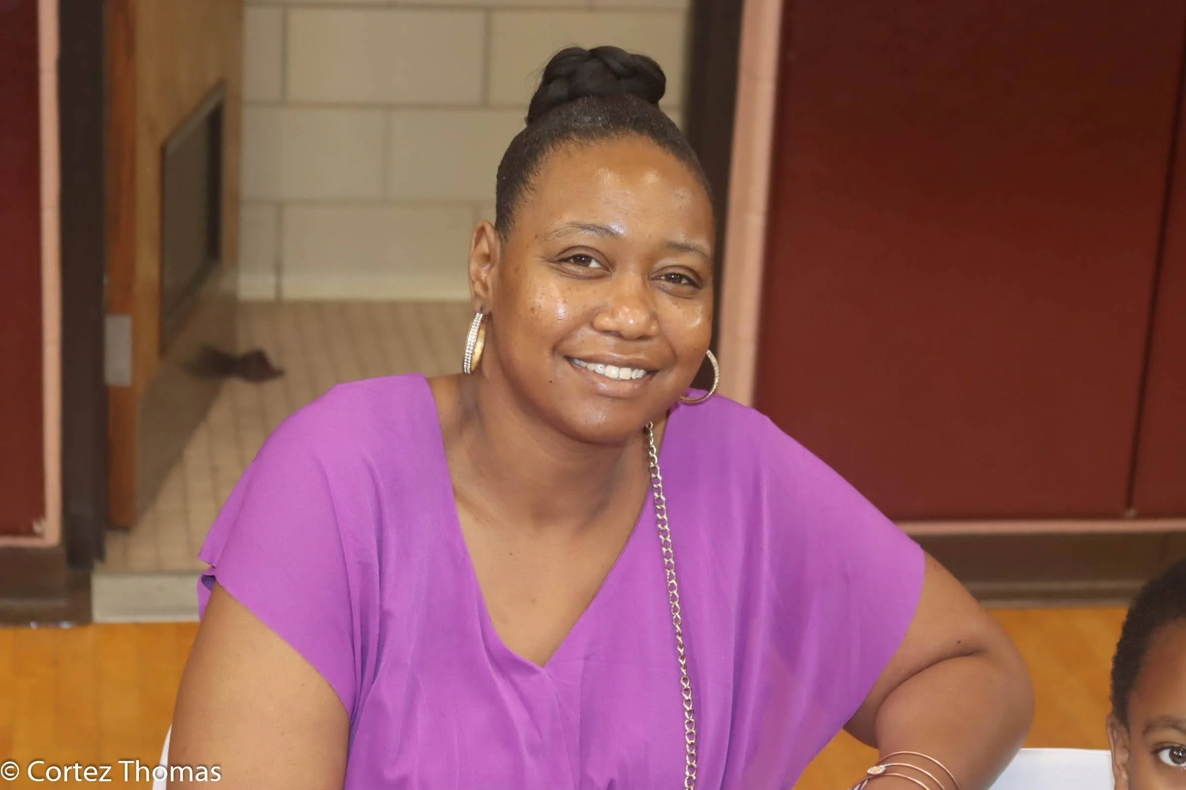 A woman with dark hair styled in a bun, wearing a purple top and gold hoop earrings, smiling at the camera, sitting at a table indoors.