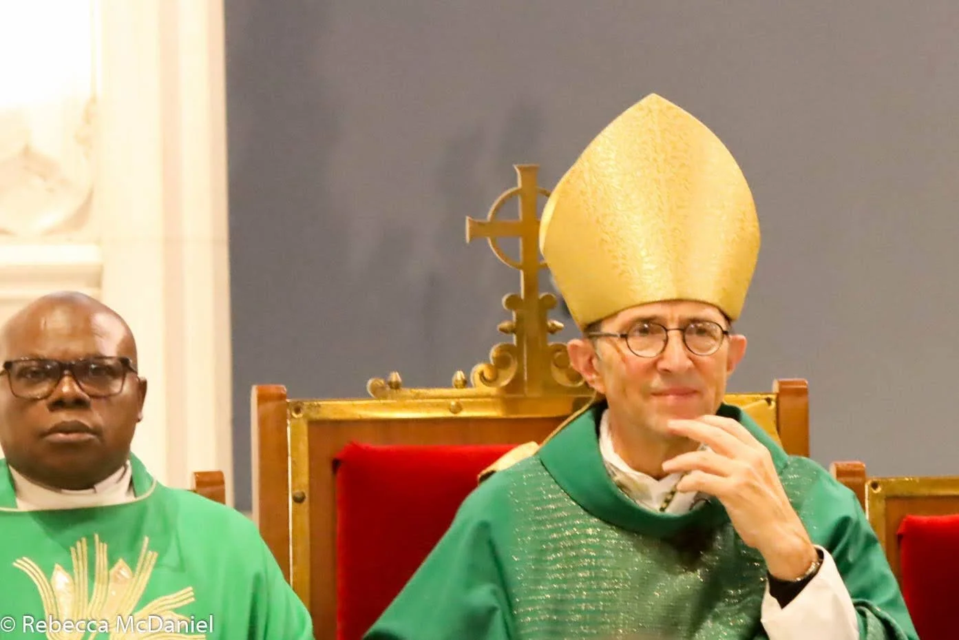 A man wearing a green religious robe and a tall gold mitre hat, seated on a throne with a red backrest, and another man in a similar green robe beside him, inside a church.