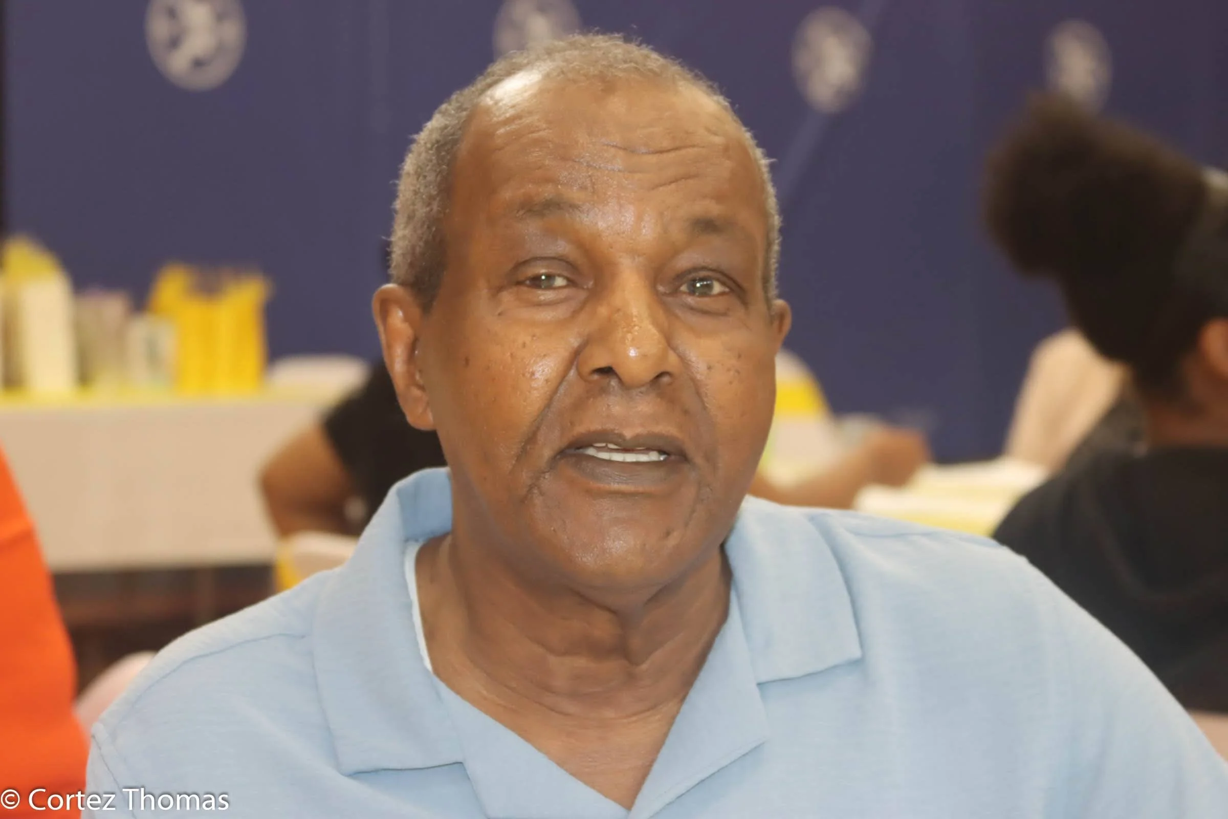 An older man with short gray hair wearing a light blue polo shirt, indoors with a blue background, possibly in a classroom or event setting.