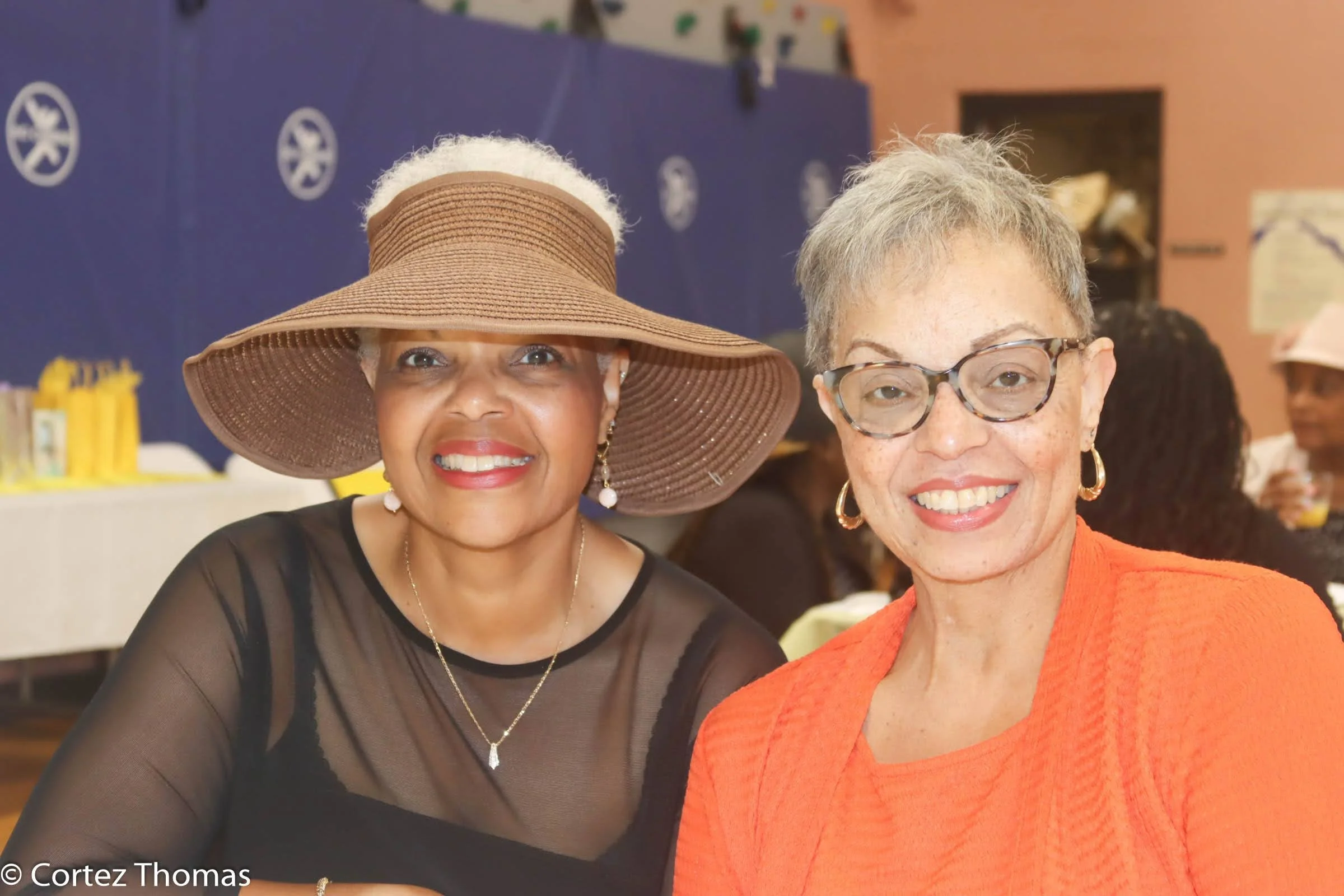 Two women smiling at a social event, one wearing a large tan sunhat and the other wearing glasses and an orange top.